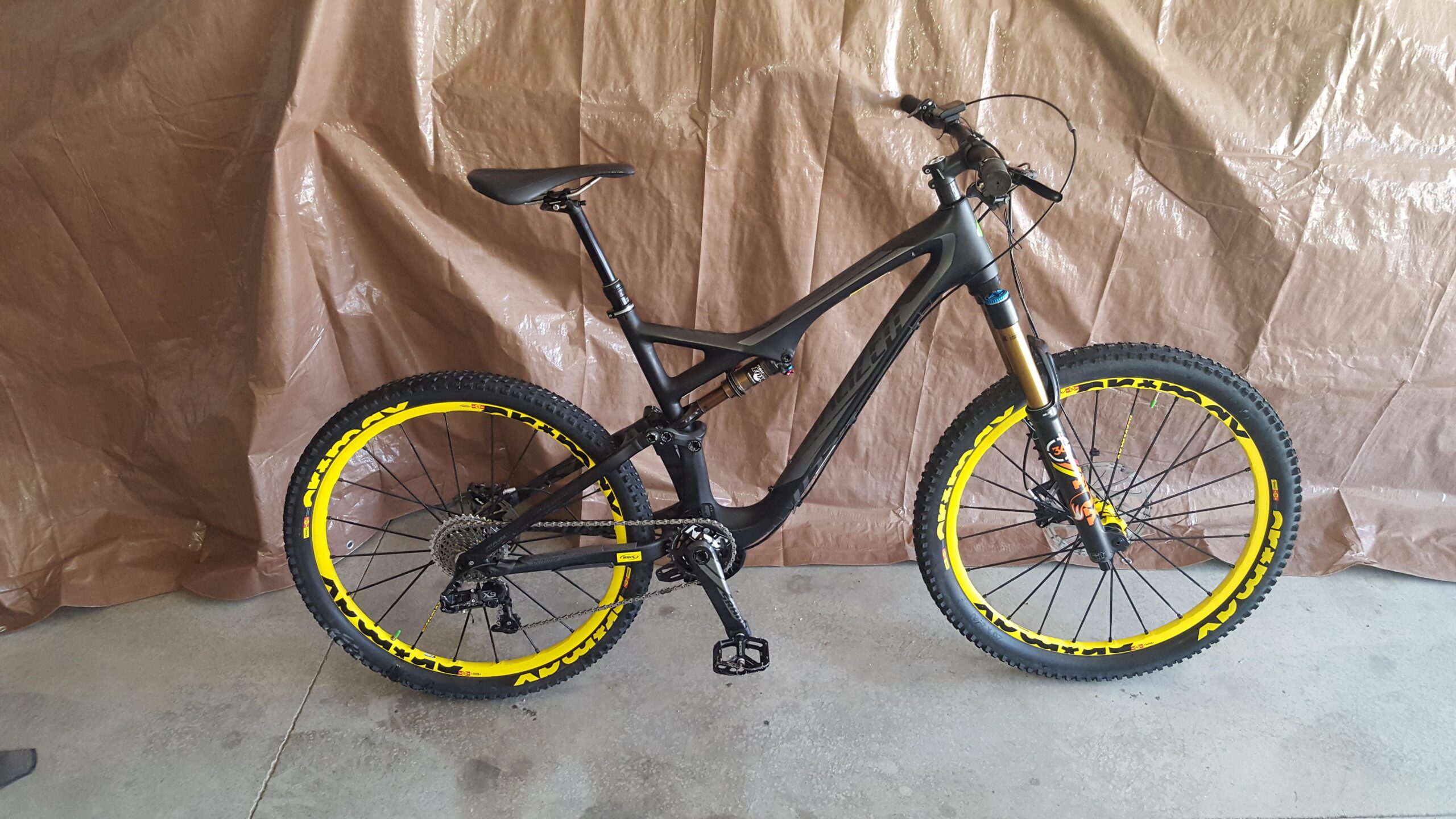 Specialized STUMPJUMPER EVO HT: A mountain bike with a black frame and bright yellow rims, featuring a suspension fork and a durable design. The bike is positioned upright on a concrete surface against a brown tarp background.