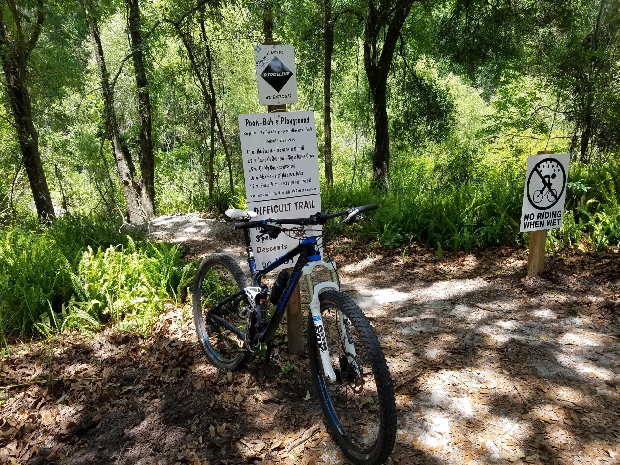 Trek Fuel EX 9.7 29: A mountain bike parked next to a trail sign indicating "Pooh Bah's Playground," which provides information about the trail's difficulty and distances to various landmarks. Surrounding the trail are lush green ferns and trees, with a secondary sign warning "No Riding When Wet." The area appears sunny and inviting for outdoor biking enthusiasts.