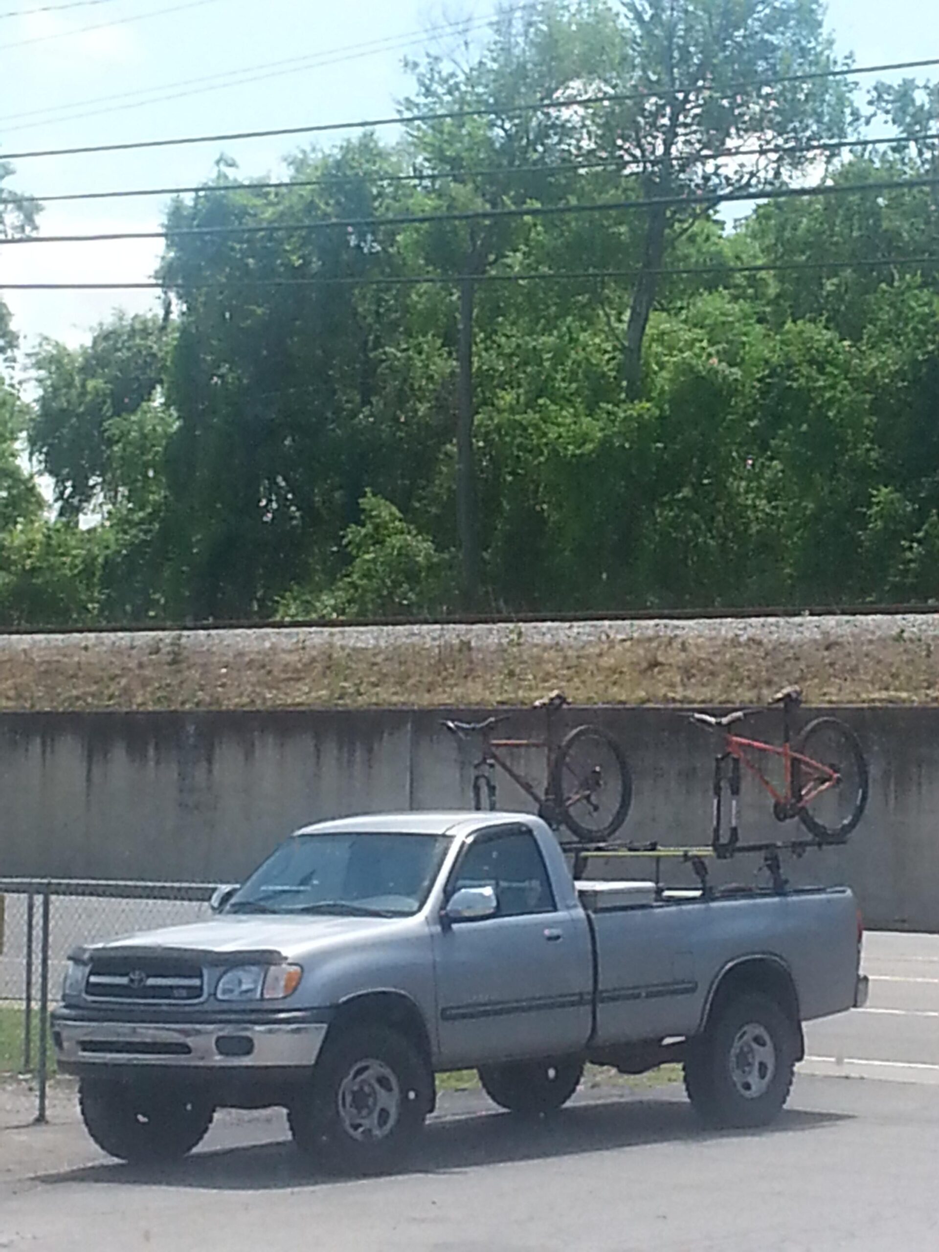 Surly Karate Monkey: A silver pickup truck parked with two bicycles secured on a bike rack in the truck bed. In the background, there are green trees and a concrete wall, along with power lines overhead. The scene is set on a sunny day.