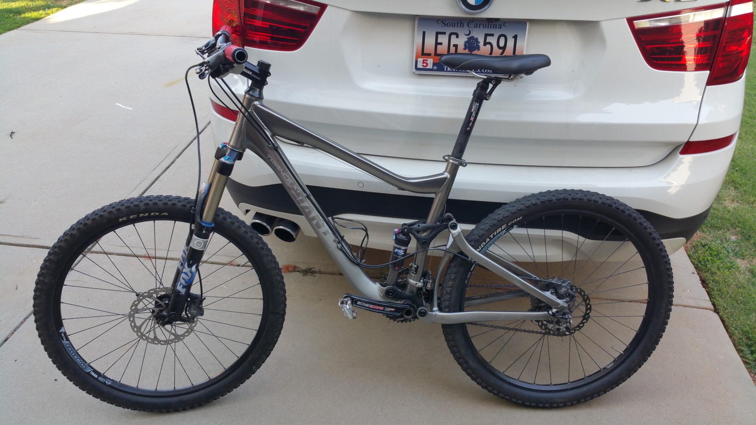 Giant Trance X1: A mountain bike with a silver frame, black tires, and front suspension is parked next to a white SUV. The bike features disc brakes and branded components, with a license plate visible on the vehicle in the background. The setting is an outdoor garage or driveway.