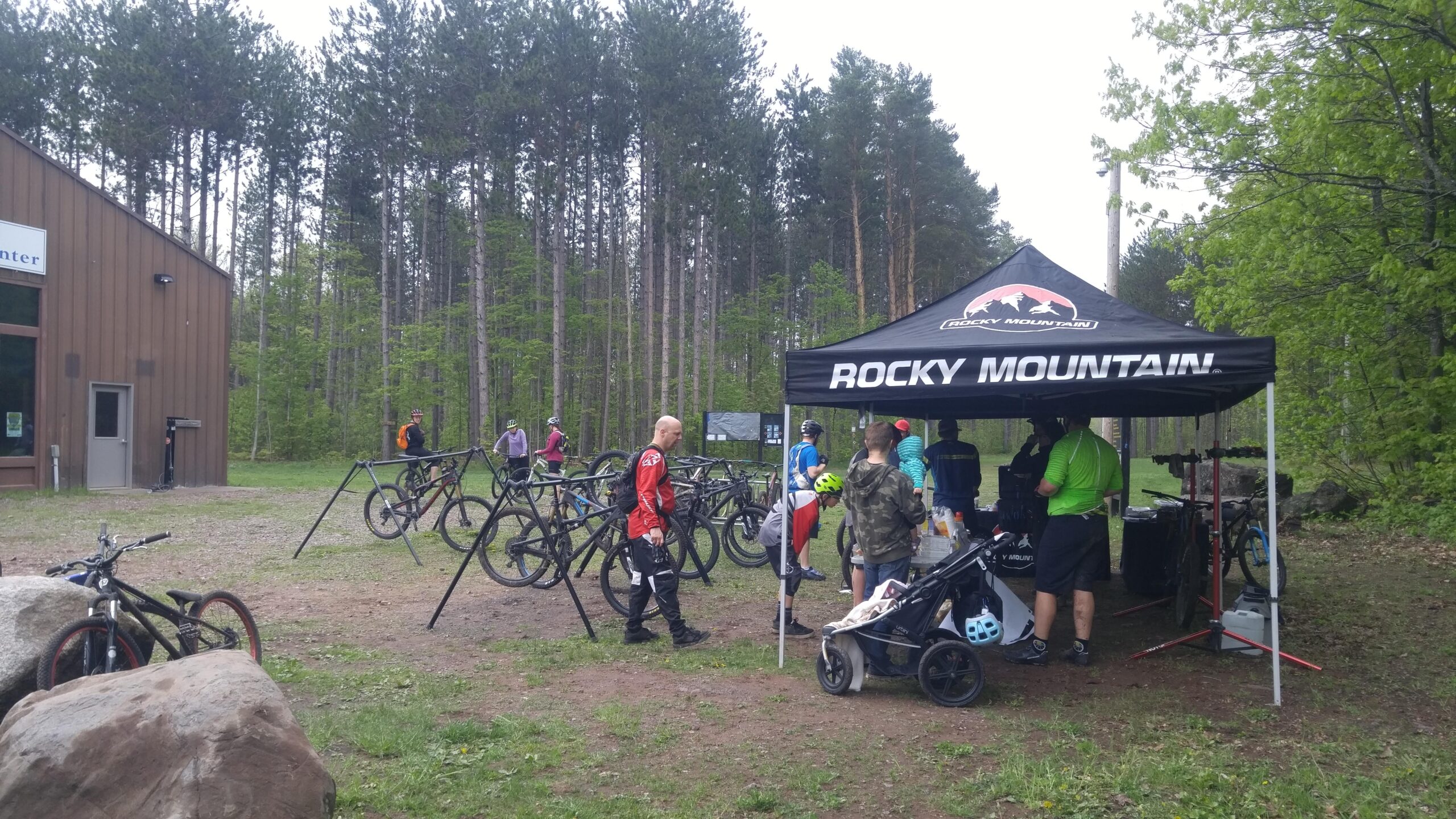 A gathering of cyclists at an outdoor event near a service center. Several mountain bikes are parked in racks, while participants engage with event staff under a branded tent. The scene is set in a forested area with green trees and a cloudy sky. Some individuals are wearing cycling gear, and a stroller is present, indicating a family-friendly atmosphere. Michigan Tech Trails mountain bike trail.