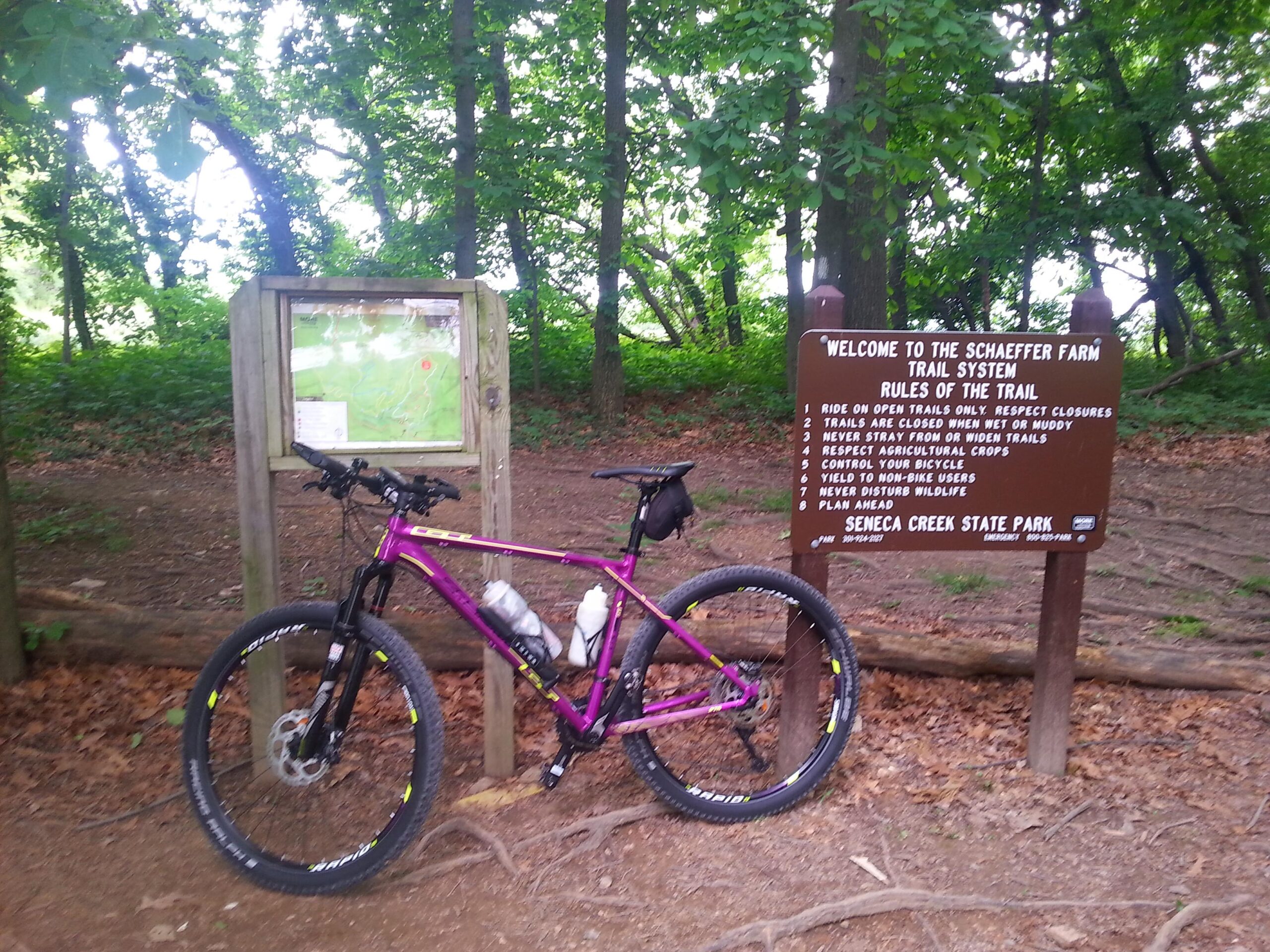 A vibrant purple mountain bike rests next to a wooden trail sign at the Schaeffer Farm Trail System in Seneca Creek State Park. The sign outlines the rules of the trail, surrounded by lush greenery and trees, indicating a natural outdoor setting. A map is visible on a board above the sign. Schaeffer Farms mountain bike trail.