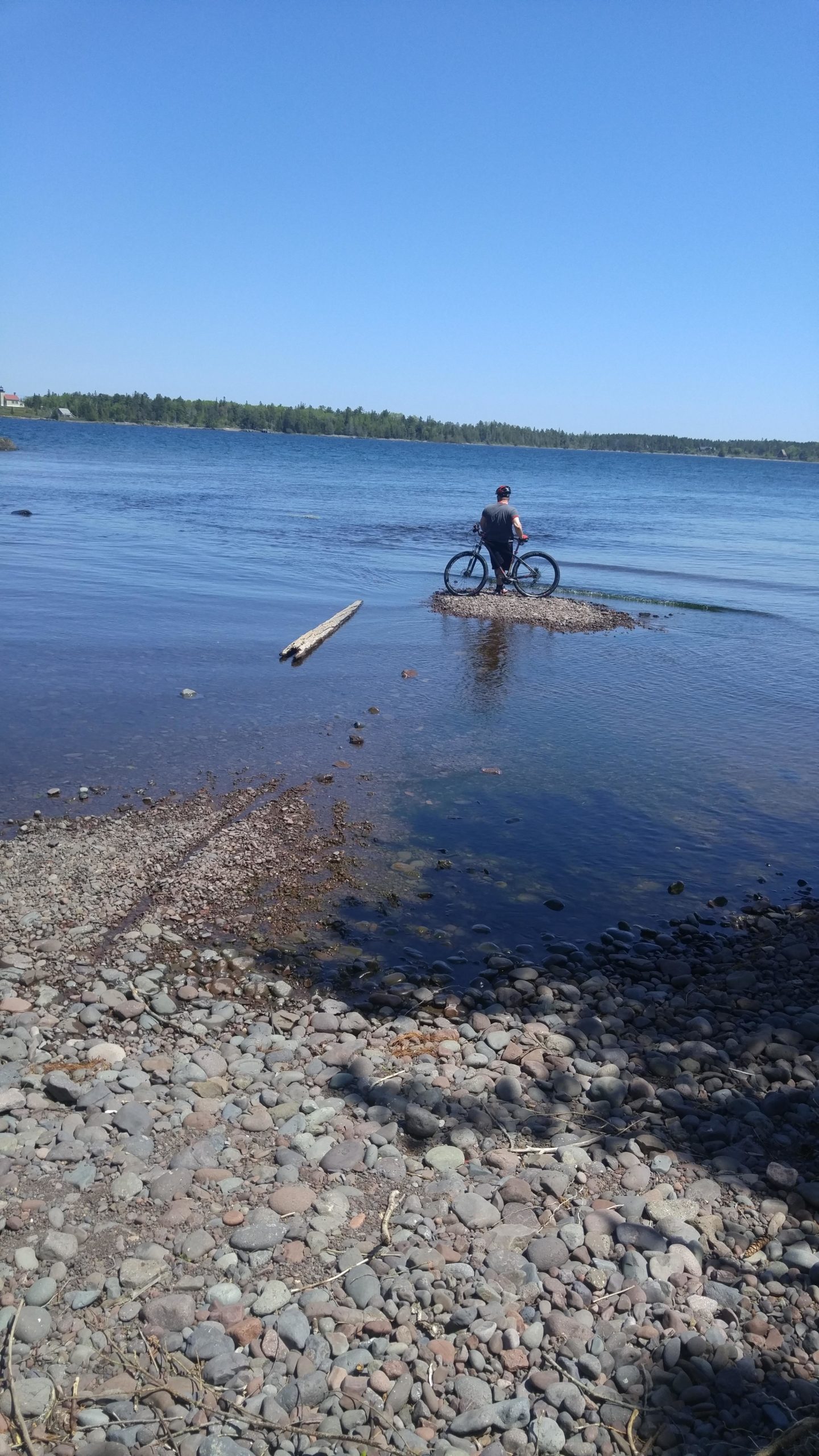 A person standing next to a bicycle on a rocky shore, partially submerged in shallow water, with a calm lake and clear blue sky in the background. Copper Harbor Trails mountain bike trail.