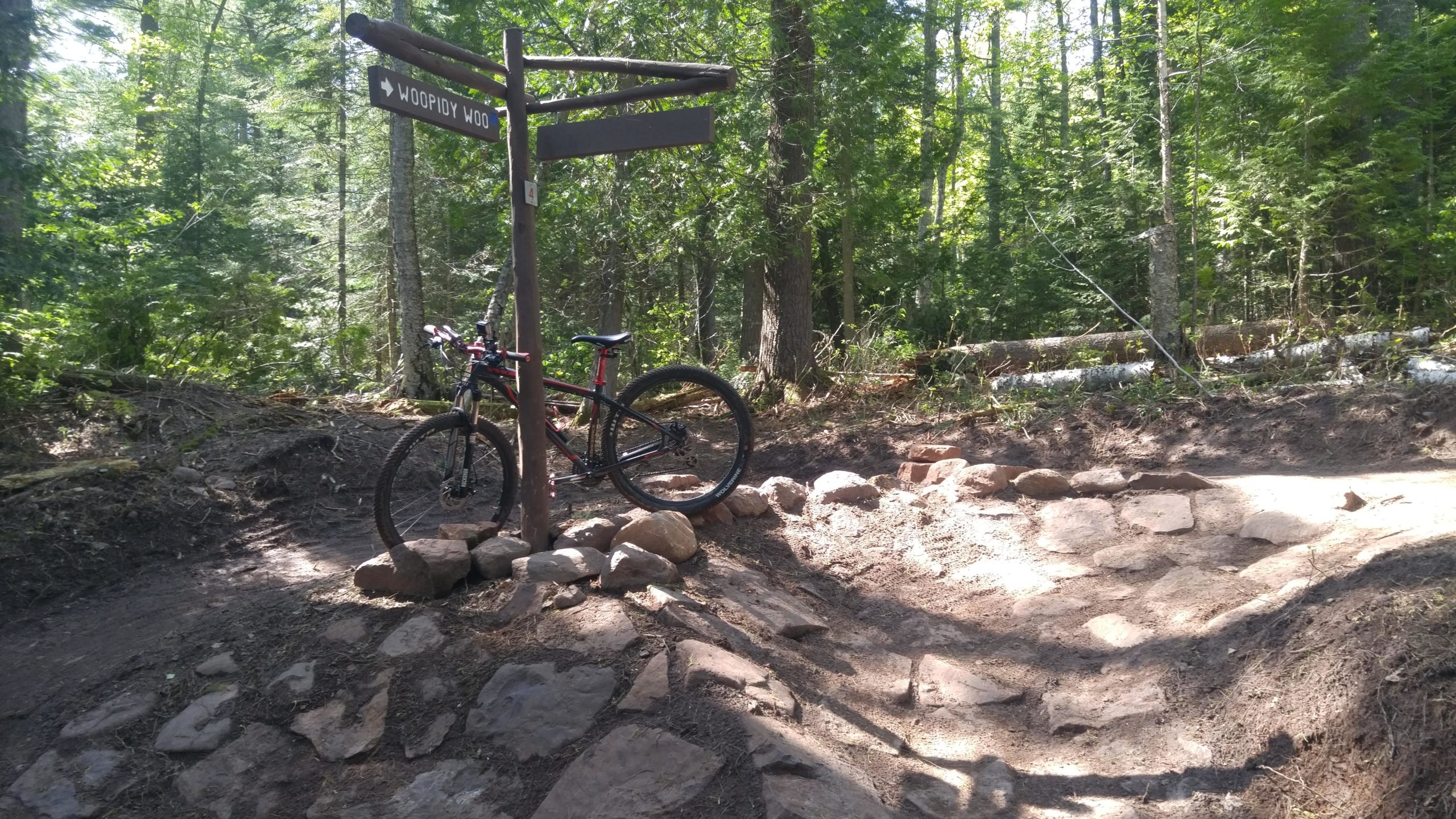 A mountain bike parked beside a wooden sign pointing towards a trail labeled "Woofidy Woo," with a rocky path leading into a lush, green forest. Copper Harbor Trails mountain bike trail.