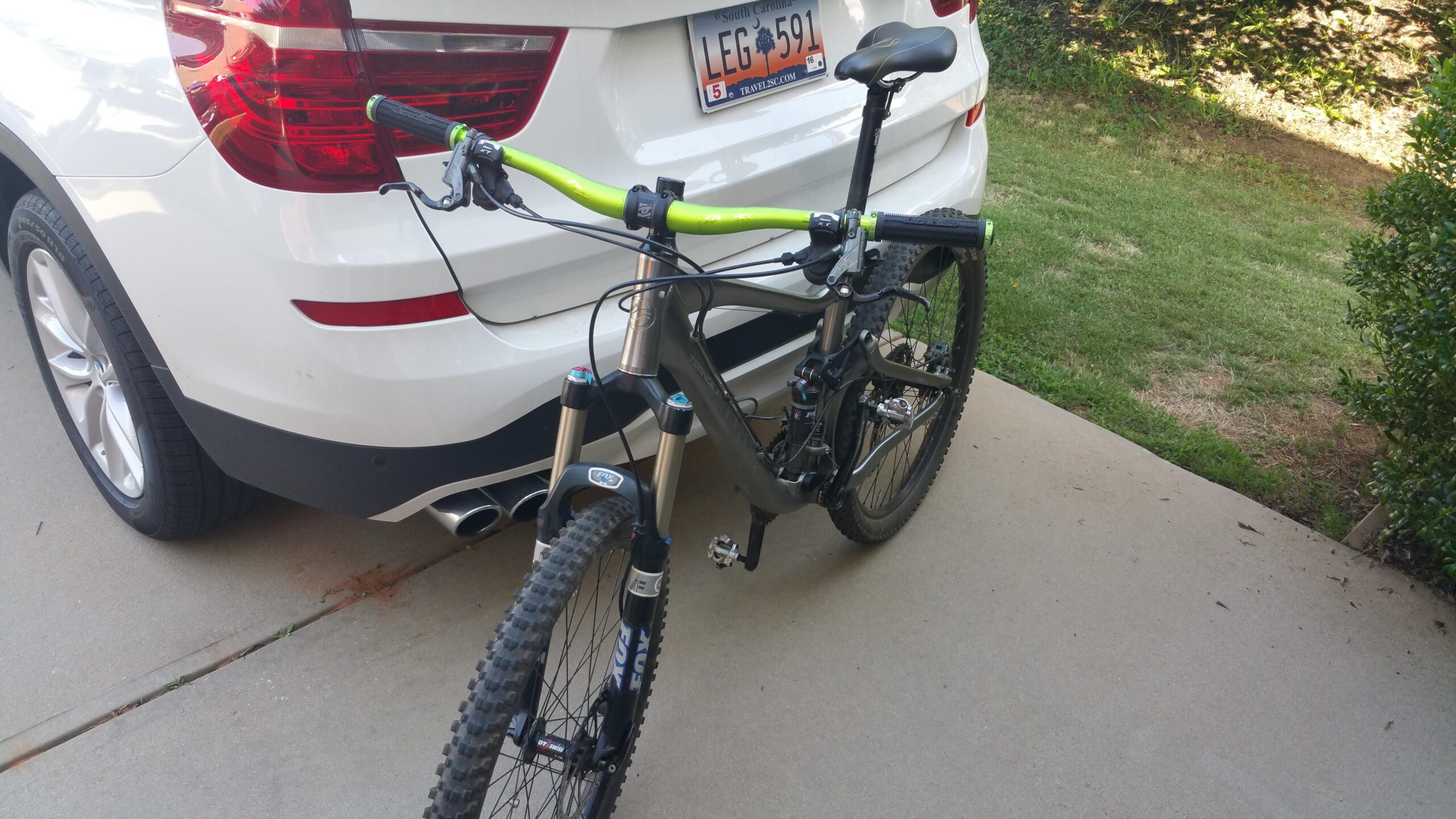 Giant Trance X1: A mountain bike with a dark frame and bright green handlebars is parked next to a white SUV in a driveway. The SUV's rear is visible, showcasing its taillights and license plate, with a patch of grass and bushes in the background.