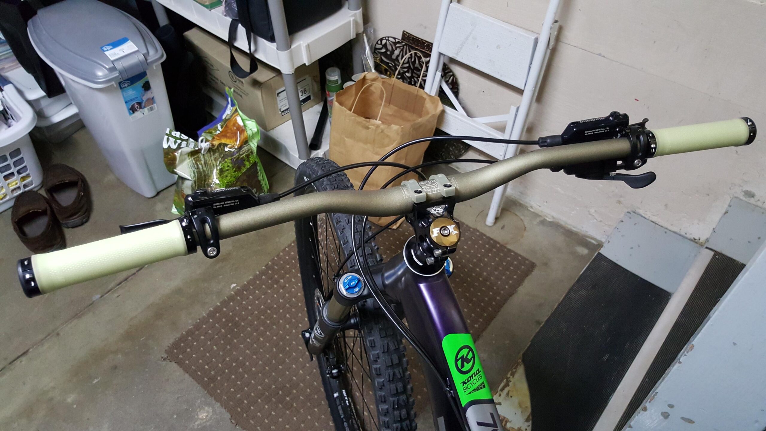 Kona Honzo: A close-up view of a bicycle's handlebars, featuring textured light green grips and a black brake lever on the right. The bike's frame is purple, and it has a gold-colored suspension piece at the top of the frame. The background shows a cluttered garage space with various items, including a trash bin, bags, and a pair of brown slippers on a concrete floor.