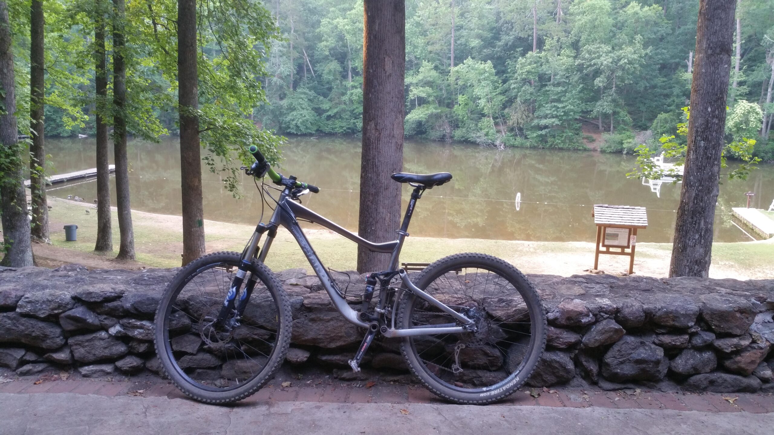 A mountain bike is leaning against a stone wall, with a serene lake and wooded area in the background. The setting is tranquil, featuring tall trees and a small dock at the edge of the water. Paris Mountain State Park mountain bike trail.