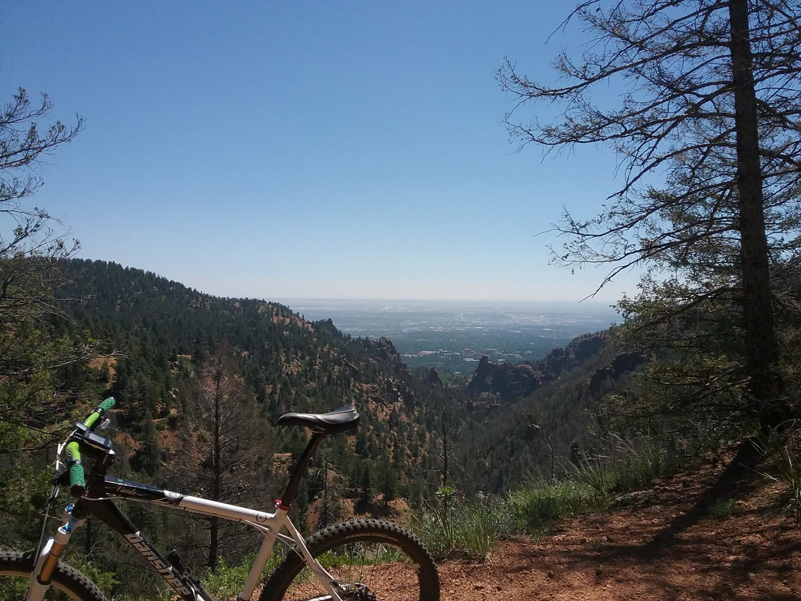 A mountain bike rests on a trail overlooking a scenic valley. Tall pine trees frame the view, which features rolling hills and a clear blue sky. The landscape showcases a vibrant mix of greenery and rocky outcrops, inviting outdoor adventure. Columbine mountain bike trail.
