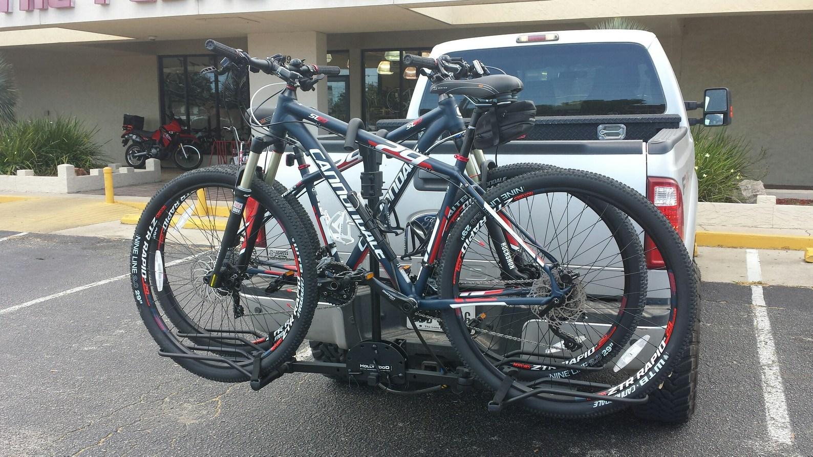 Cannondale Trail SL 29 2: Two mountain bikes are mounted on a bike rack attached to the back of a silver pickup truck. The bikes are in a parking lot, with a building visible in the background, along with some greenery and another vehicle. The scene conveys an outdoor, adventurous atmosphere.
