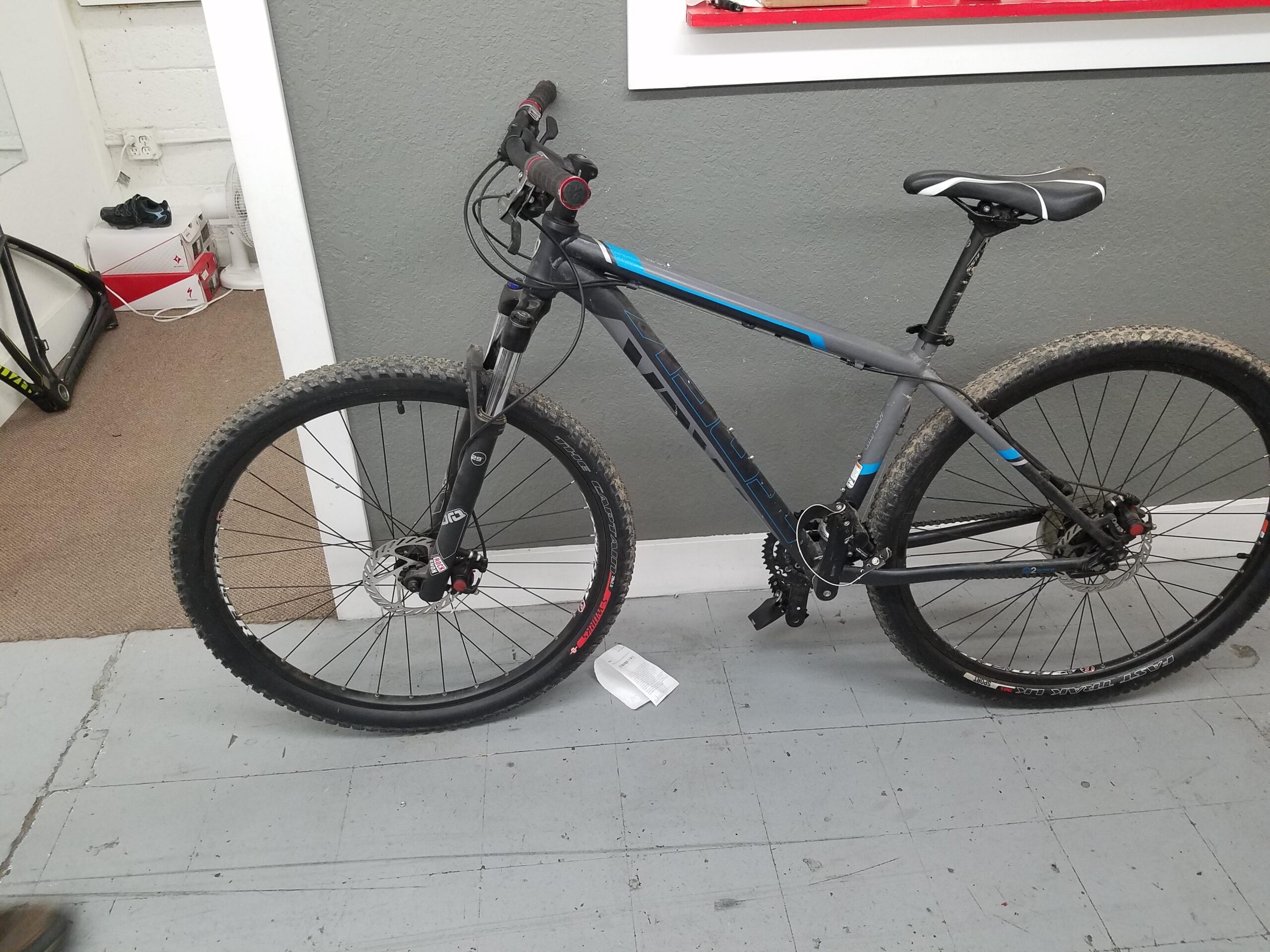 Trek Mamba: Image of a gray mountain bike with blue accents, placed in an indoor setting. The bike features knobby tires, a front suspension fork, and a textured seat. In the background, there are hints of additional bike parts and a fan, with a gray wall and light-colored floor.