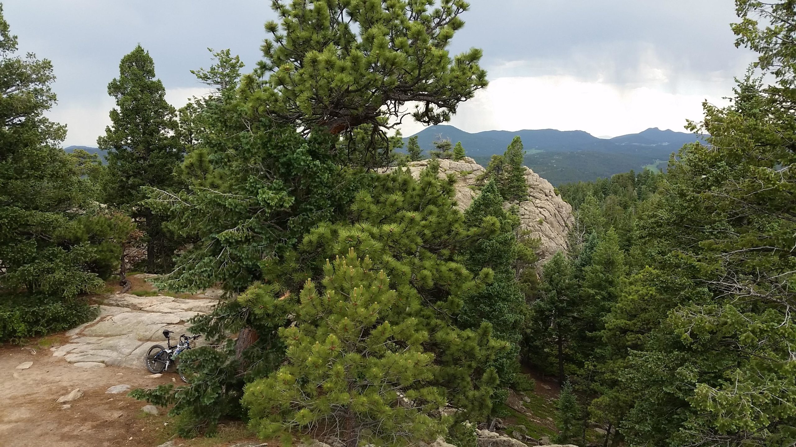 A scenic view of a rocky landscape surrounded by lush green pine trees, with a mountain bike resting against a boulder. The sky is partly cloudy, with distant mountains visible in the background. 3 Sisters / Alderfer mountain bike trail.