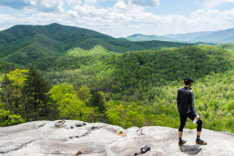 A person stands on a rocky outcrop overlooking a lush green valley surrounded by rolling hills under a partly cloudy sky. The individual is wearing cycling gear and holding a water bottle, enjoying the scenic view of the expansive landscape.