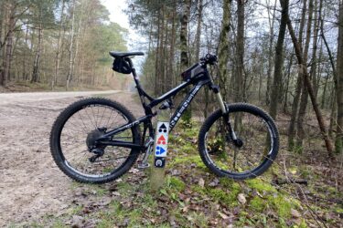 A mountain bike leaning against a wooden post in a forested area, with a dirt path visible in the background. The bike features colorful stickers on the frame and is positioned amidst fallen leaves and grass. Appelscha Rood-Blauw-Rood mountain bike trail.