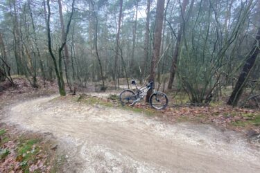 A mountain bike resting on a winding dirt trail surrounded by trees in a foggy forest setting. The trail is partially covered with leaves and has a soft, sandy surface. Appelscha Rood-Blauw-Rood mountain bike trail.