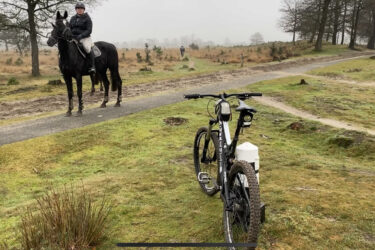 A mountain bike rests on grassy terrain in the foreground, while a horse and rider are seen in the background on a misty day. The path is surrounded by sparse trees and brush, with another rider visible in the distance. Appelscha Rood-Blauw-Rood mountain bike trail.