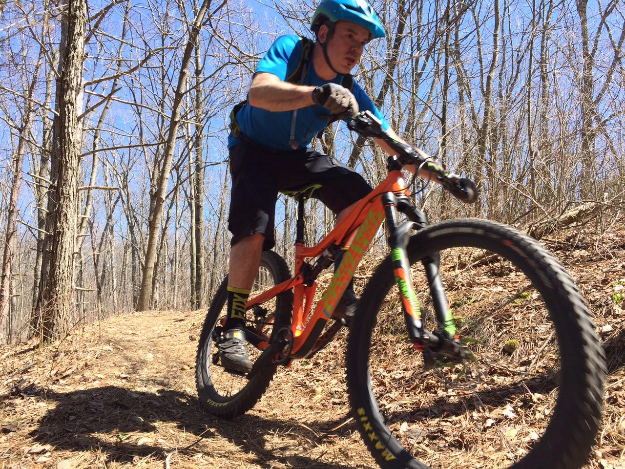 Santa Cruz 5010: A mountain biker in a blue shirt and helmet is riding an orange bike on a narrow trail surrounded by bare trees. The cyclist is leaning forward, demonstrating speed and agility as they navigate the terrain on a sunny day.