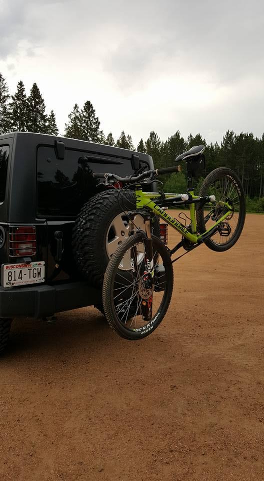 A black vehicle parked on a dirt surface with a mountain bike mounted on the rear. The bike is bright green and secured to the back of the vehicle, which has a spare tire attached. In the background, there are trees and an overcast sky. Nine Mile mountain bike trail.