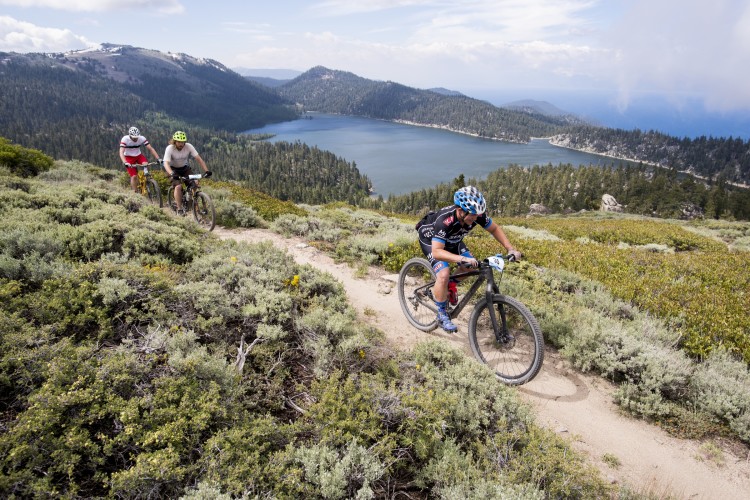 Three mountain bikers navigating a trail on a hillside, surrounded by lush greenery and overlooking a serene lake in the background. The scene captures the beauty of outdoor cycling in a mountainous landscape with distant hills and a partly cloudy sky.