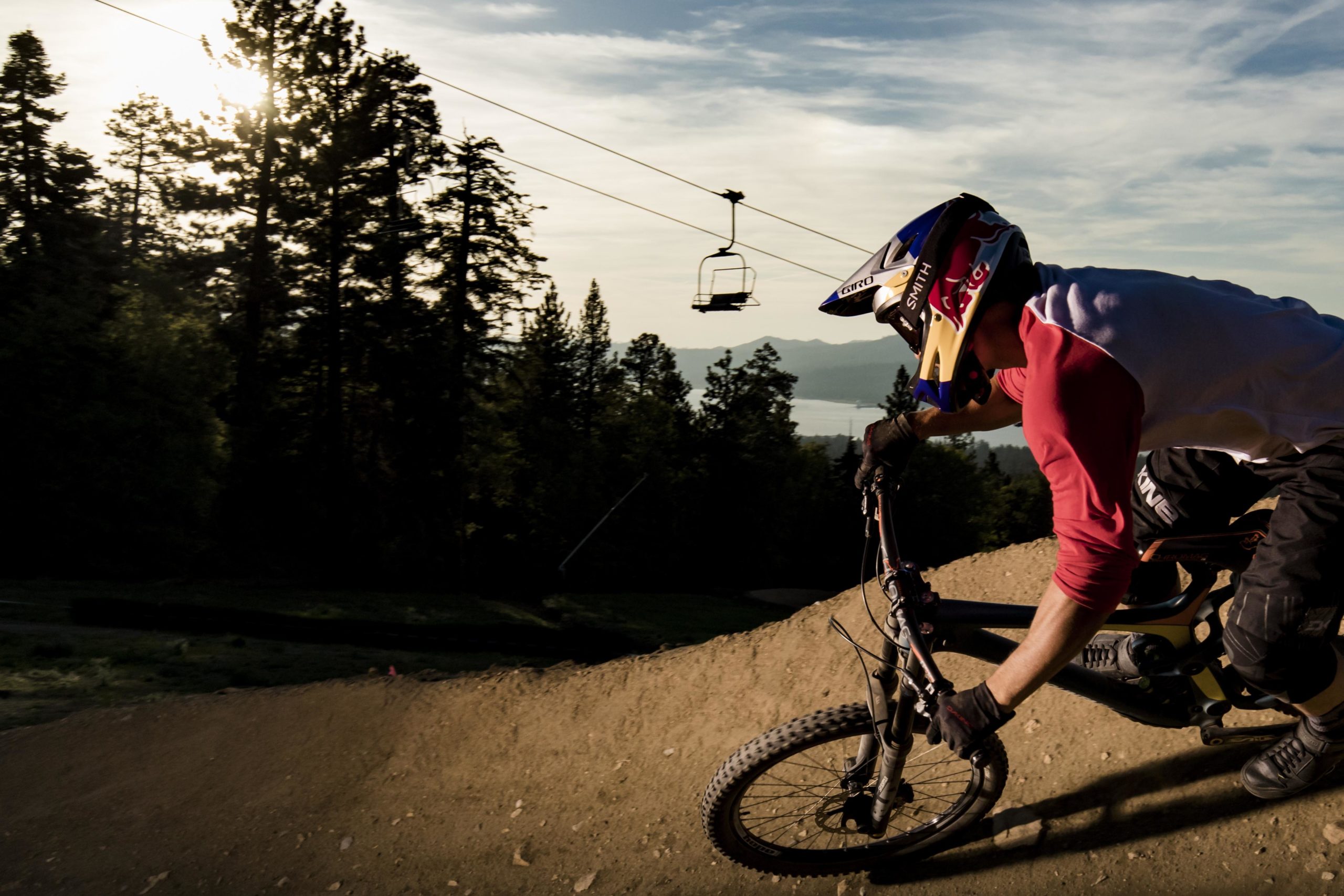 A mountain biker in a helmet and protective gear leans into a turn on a dirt trail, surrounded by tall trees and a scenic backdrop of a lake and mountains. A ski lift can be seen in the background, suggesting a mountainous terrain ideal for biking. The sun is low in the sky, creating a dramatic lighting effect. Big Bear Mountain Resort mountain bike trail.