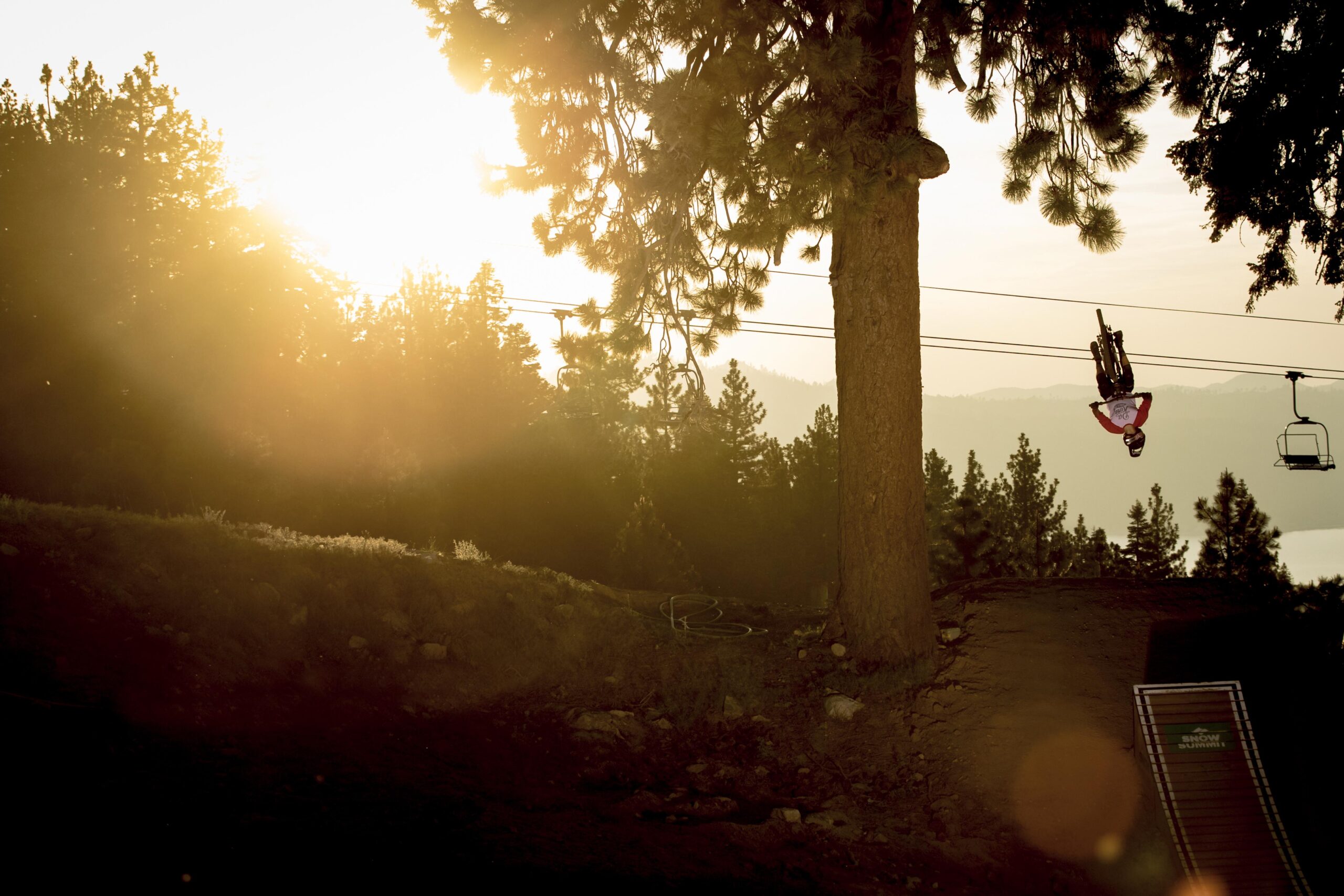 A mountain biker performs a trick mid-air against a backdrop of a sunset, with a ski lift visible in the background and tall trees surrounding the scene. The sun casts a warm glow, highlighting the rider's dynamic pose and the natural beauty of the landscape. Big Bear Mountain Resort mountain bike trail.