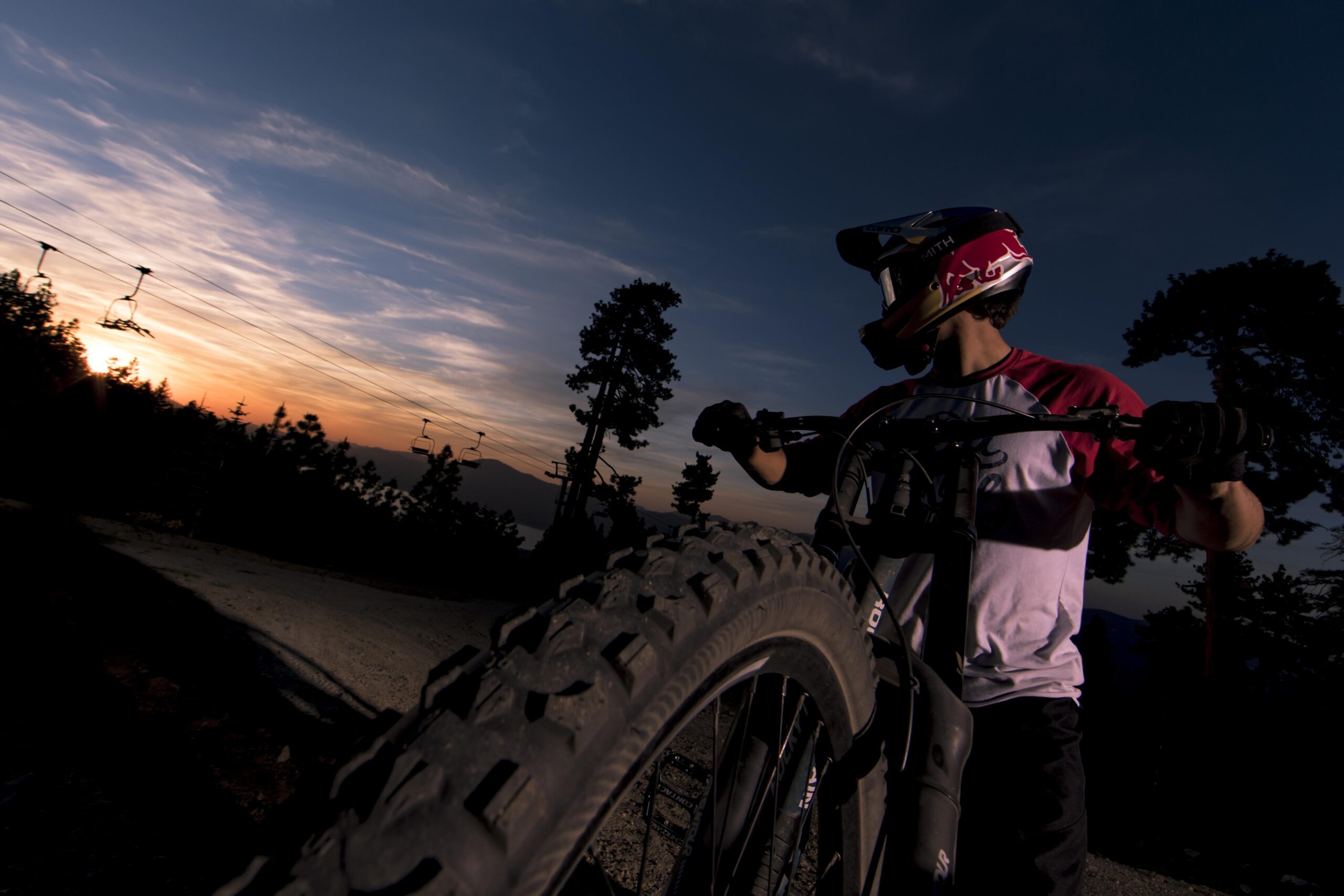 A mountain biker in a helmet and jersey stands beside their bike, silhouetted against a vibrant sunset. The image captures the bike's tire prominently in the foreground and features ski lift cables in the background, surrounded by trees and mountains. The sky is painted with hues of orange, pink, and blue, creating a dramatic backdrop. Big Bear Mountain Resort mountain bike trail.