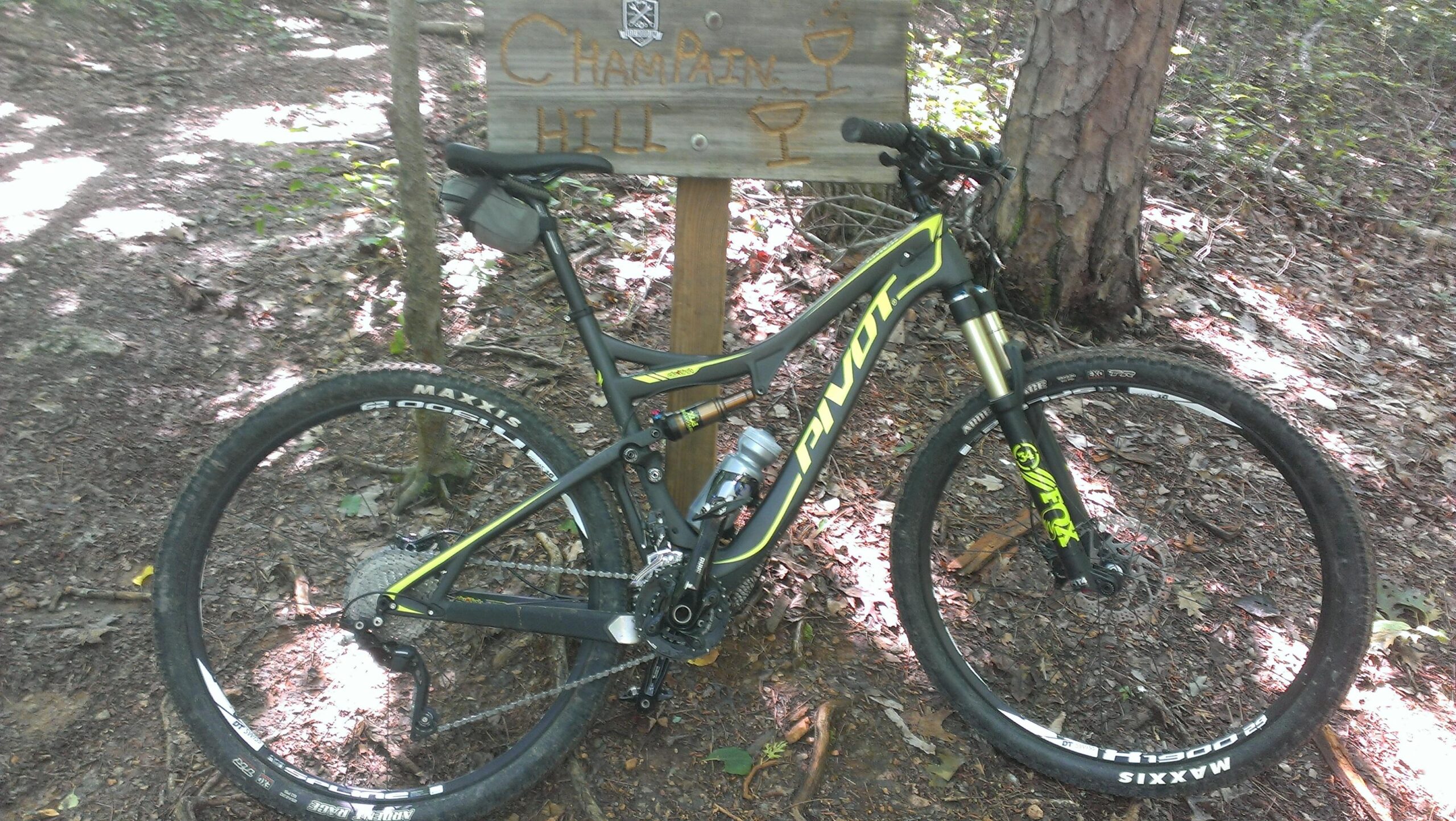 A mountain bike is parked next to a wooden sign that reads "Champaign Hill" in a forested area. The bike features a black and yellow frame and is positioned on a dirt trail with scattered leaves and sunlight filtering through the trees in the background. Chicopee Woods mountain bike trail.