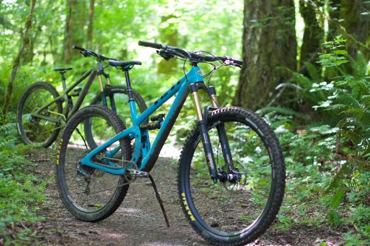 Two mountain bikes parked on a gravel trail surrounded by lush green foliage and trees in a forest setting. The bike in the foreground is bright blue, while the one in the background is a muted green. Sunlight filters through the leaves, creating a dappled light effect on the ground.