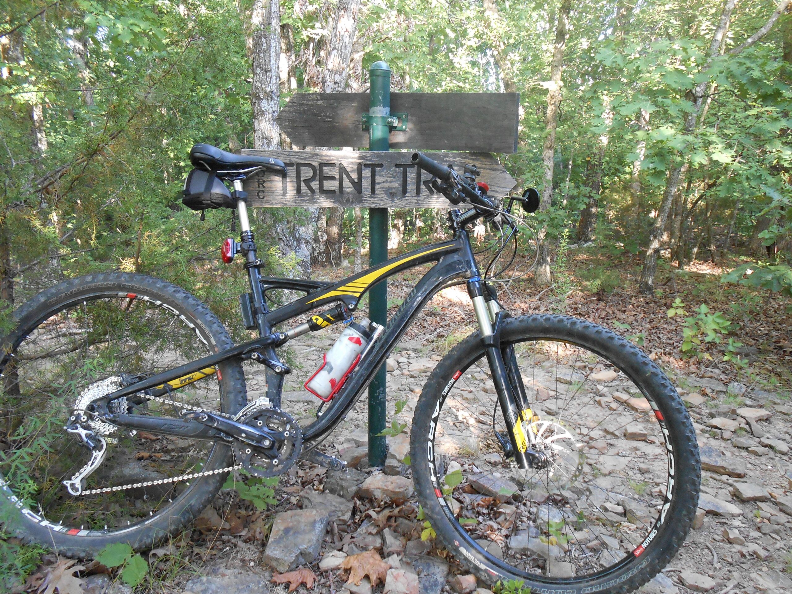 A mountain bike is parked next to a wooden signpost indicating "Trent Trail," surrounded by lush greenery and rocky terrain. Mt Kessler mountain bike trail.