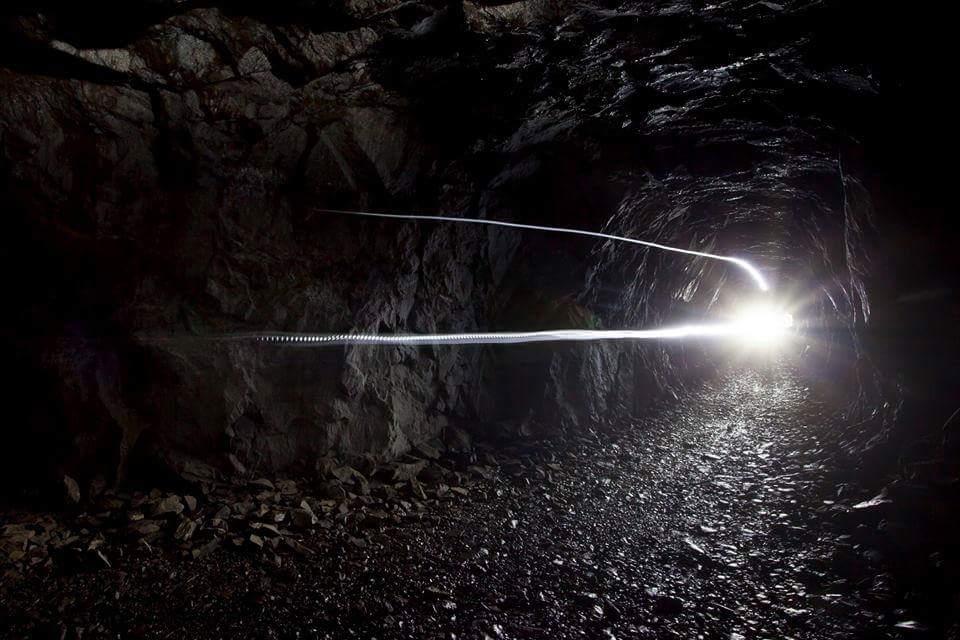 A dimly lit underground tunnel with rocky walls, featuring a bright light source at the end that creates a contrast against the darkness. The ground is uneven and scattered with stones, hinting at the rugged nature of the environment. Adventure Trails mountain bike trail.