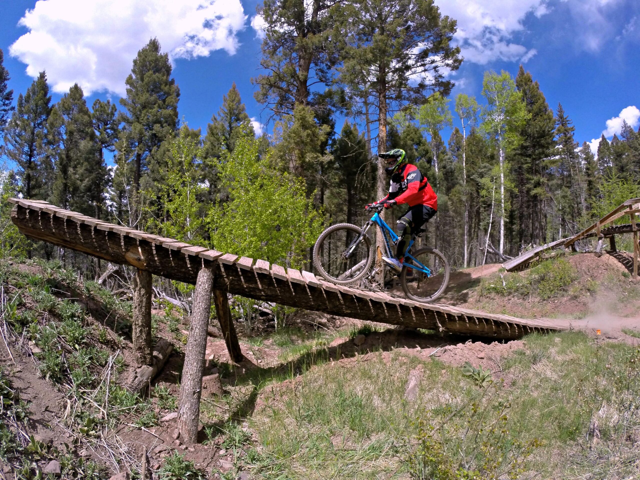 A mountain biker navigating a wooden bridge on a dirt trail, surrounded by dense trees and a clear blue sky with scattered clouds. The biker is wearing protective gear and is captured mid-air as they ride over the bridge. Angel Fire Bike Park mountain bike trail.