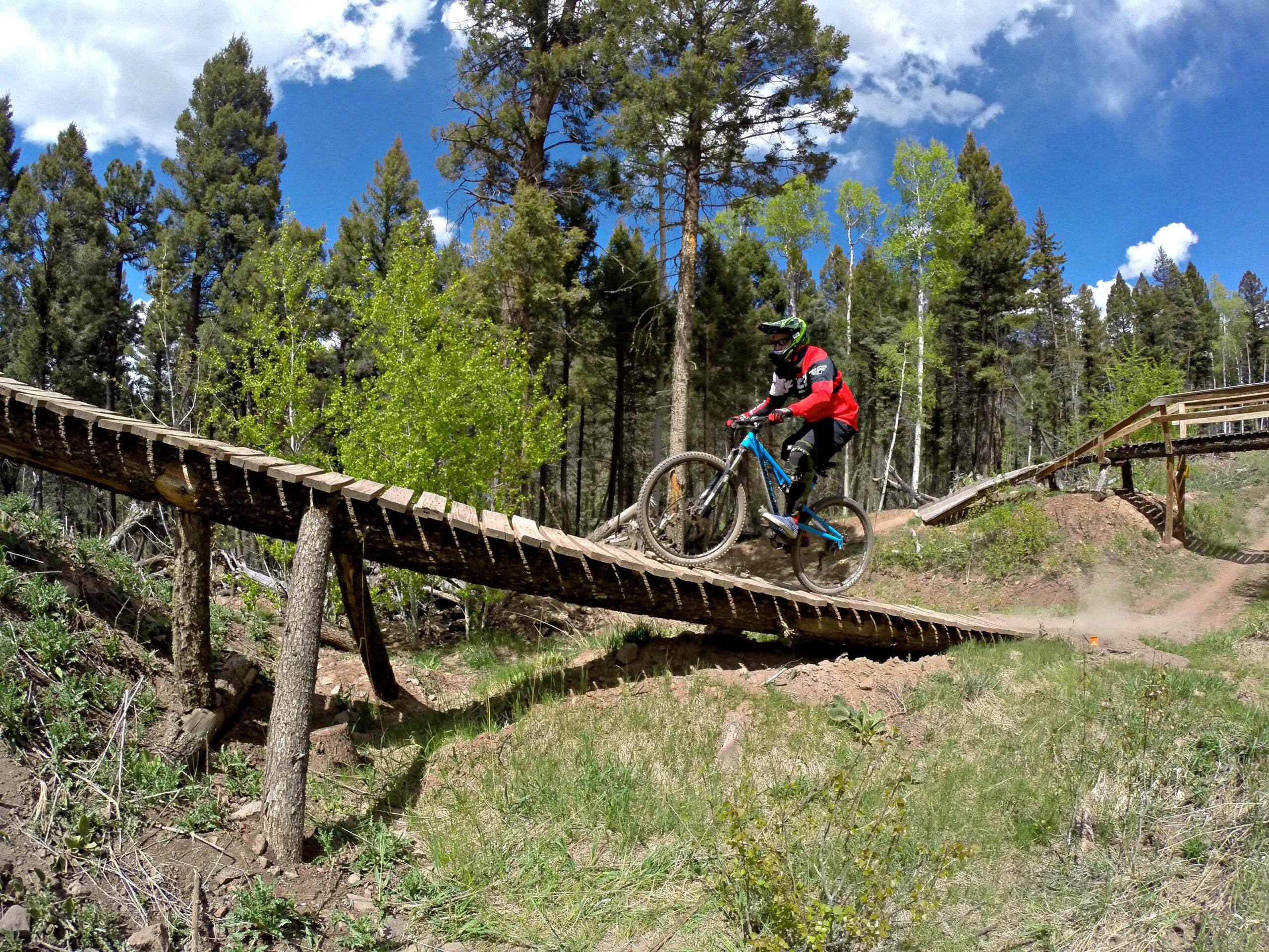 A mountain biker in a red and black outfit rides over a wooden ramp in a forested area, with green trees and blue sky in the background. The biker is captured mid-air, showcasing skillful balance and the excitement of mountain biking on a trail. Angel Fire Bike Park mountain bike trail.