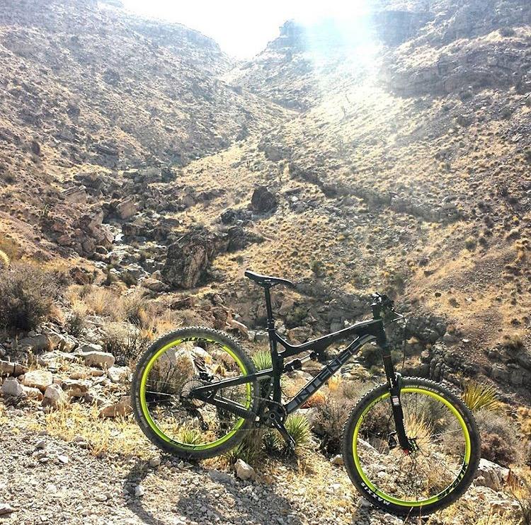 Intense Tracer 275c: A black mountain bike with green accents is parked on rocky terrain, surrounded by a dry, rugged landscape with hills and sparse vegetation in the background. Sunlight filters through, illuminating the scene.
