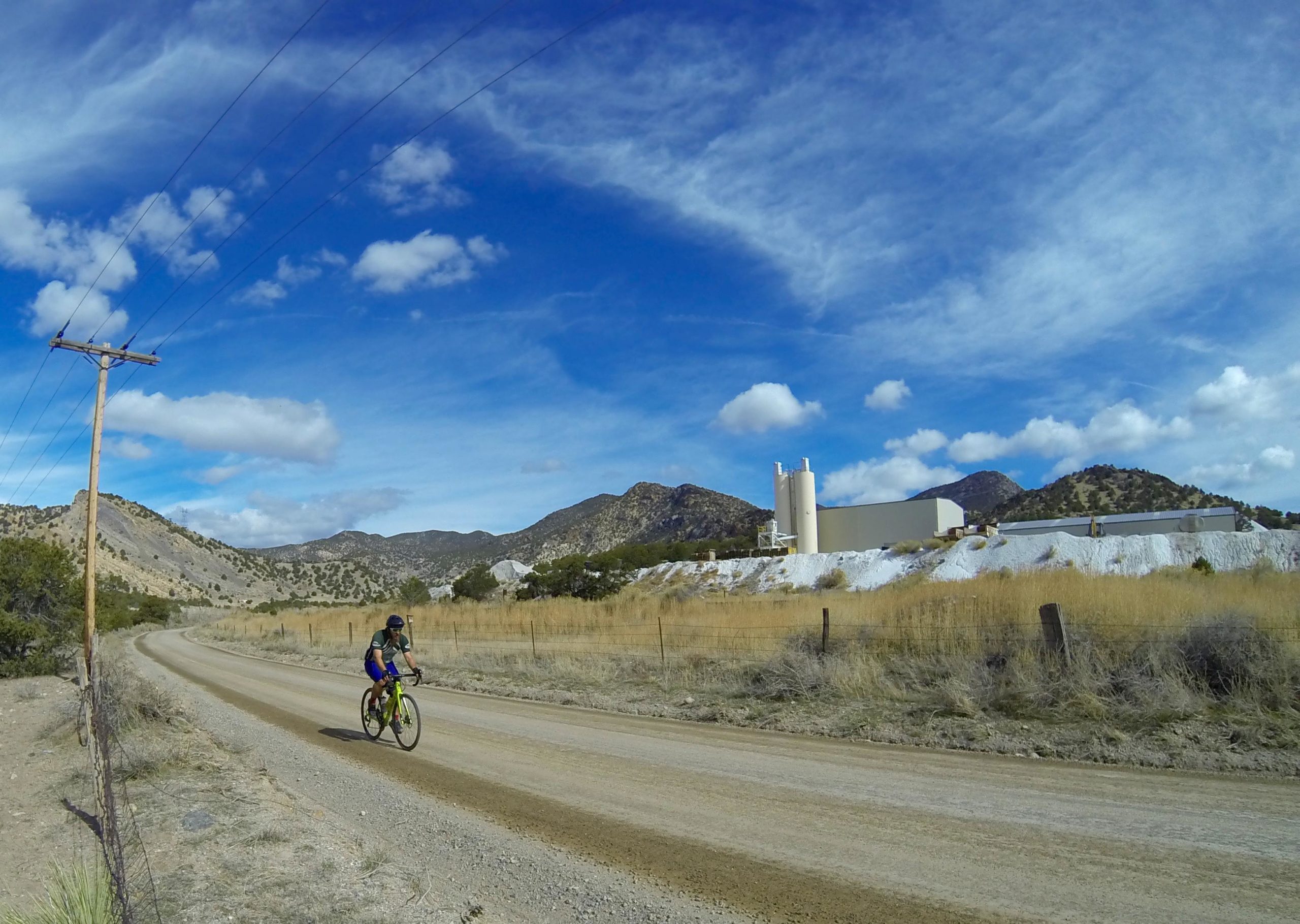 A cyclist riding a bicycle on a gravel road surrounded by dry grass and hills, with a cloudy blue sky overhead. In the background, a large white industrial building and silos can be seen against the landscape. Power lines run along the side of the road. Road #7 mountain bike trail.