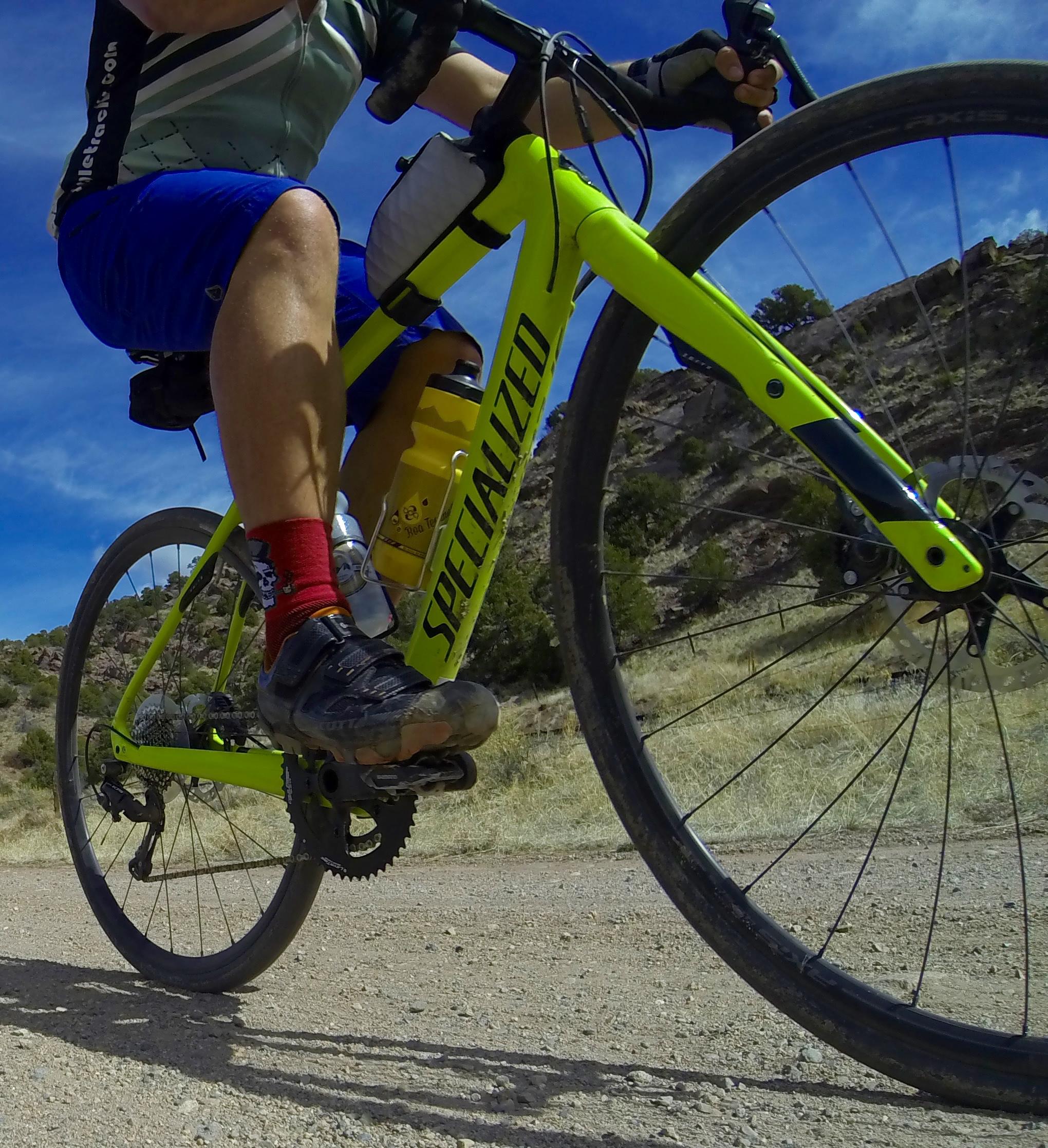 A cyclist in motion on a dirt trail, riding a bright yellow Specialized bicycle. The rider wears a light gray and green jersey, blue shorts, and red socks, showcasing a water bottle attached to the bike frame. The background features a rugged landscape with dry grass and rocky terrain under a blue sky. Road #45 mountain bike trail.