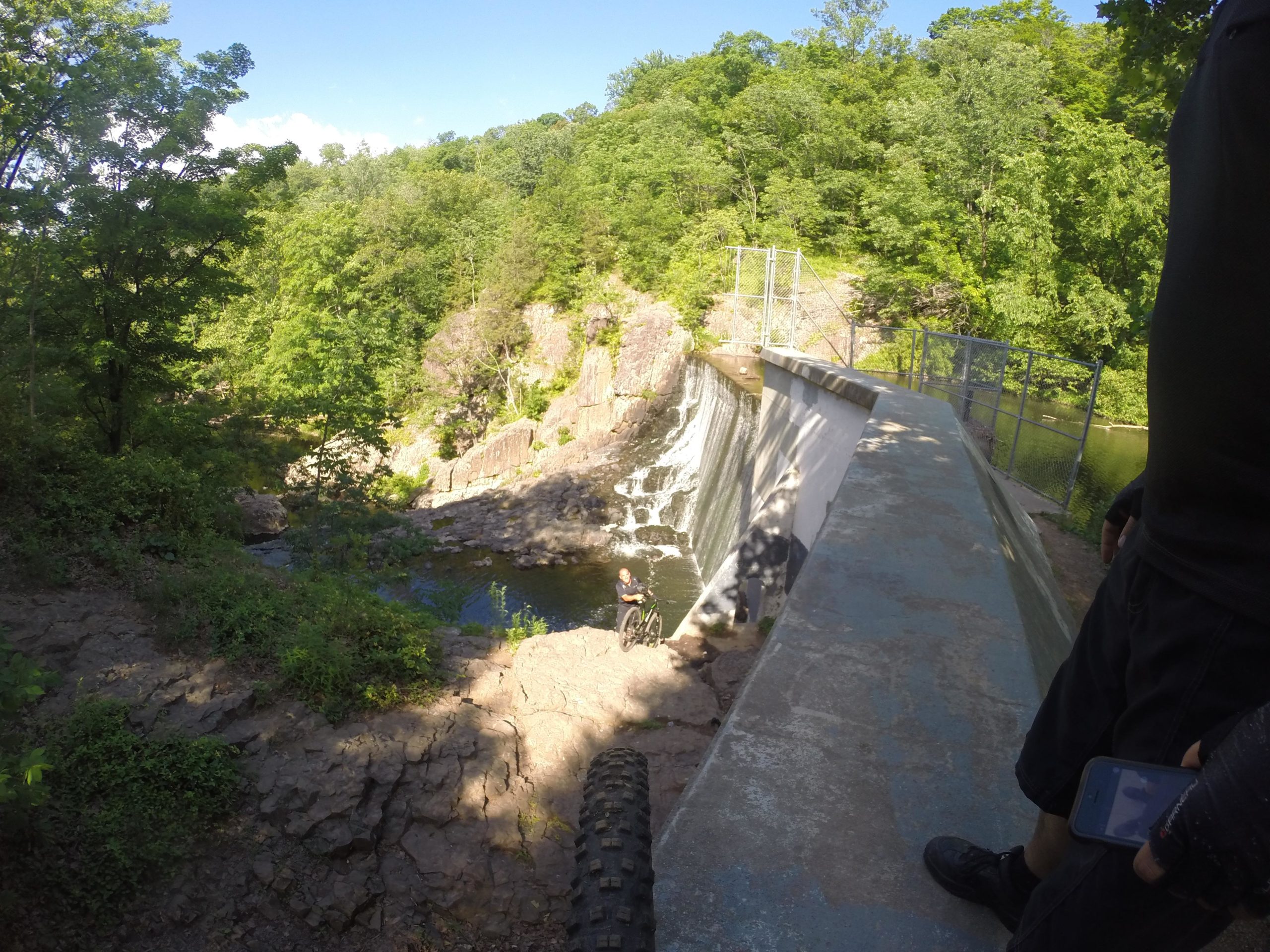 A scenic view of a waterfall cascading over a dam in a wooded area. In the foreground, a person stands near the edge of the dam, overlooking the water below. Lush green trees surround the scene, creating a tranquil natural setting on a sunny day. A rocky terrain is visible in the lower part of the image, adding to the landscape’s rugged charm. Washington Valley mountain bike trail.