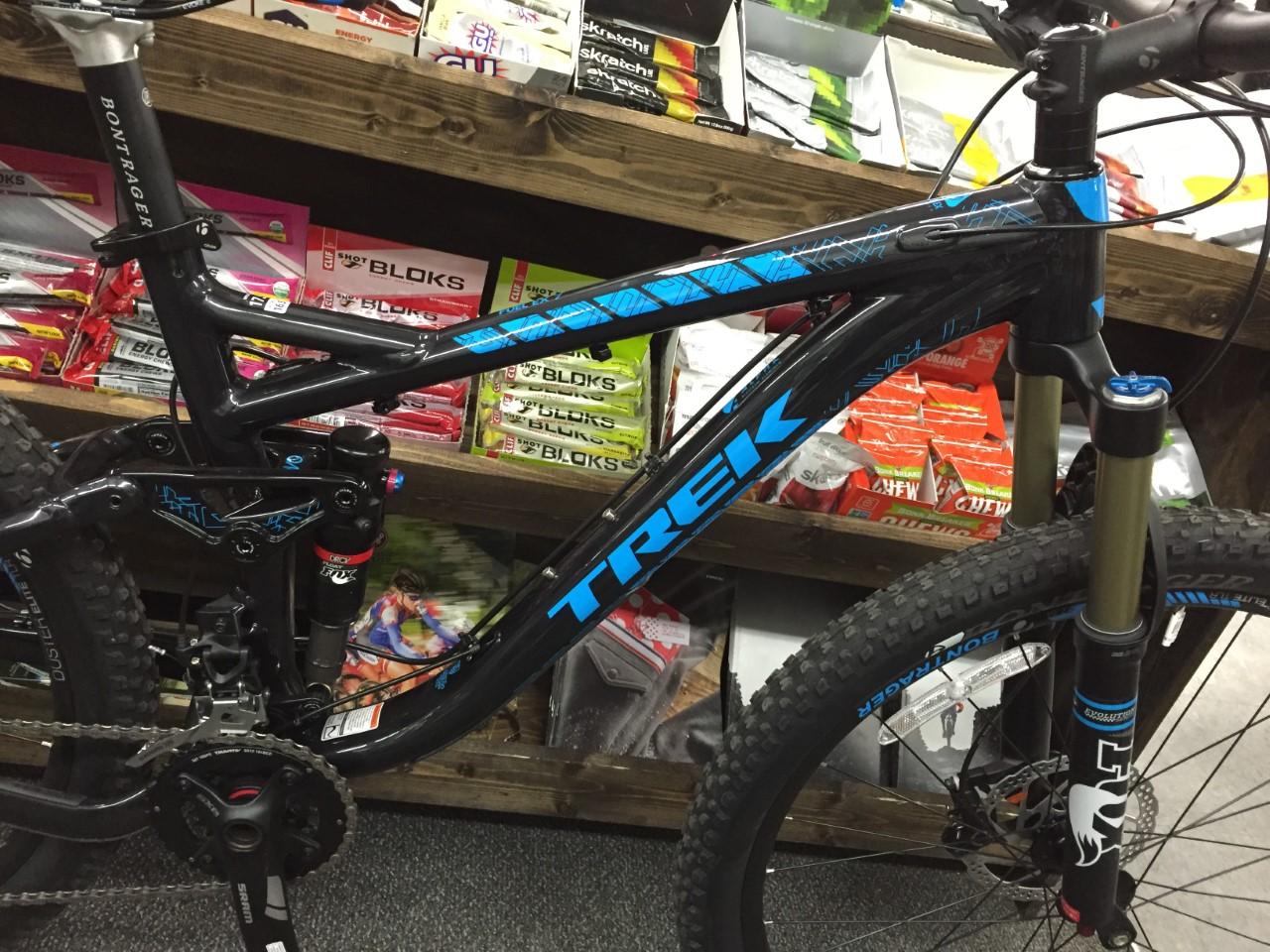 Trek Fuel EX 7: Alt text: Close-up of a black and blue Trek mountain bike displayed in a shop, with various energy snacks and products visible in the background.