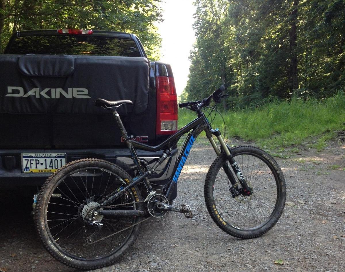 Santa Cruz Bronson: A black mountain bike leaning against the back of a truck parked on a gravel trail, surrounded by greenery. The truck has a cover with the logo "DAKINE" visible. The scene suggests an outdoor adventure setting, likely for biking or hiking.