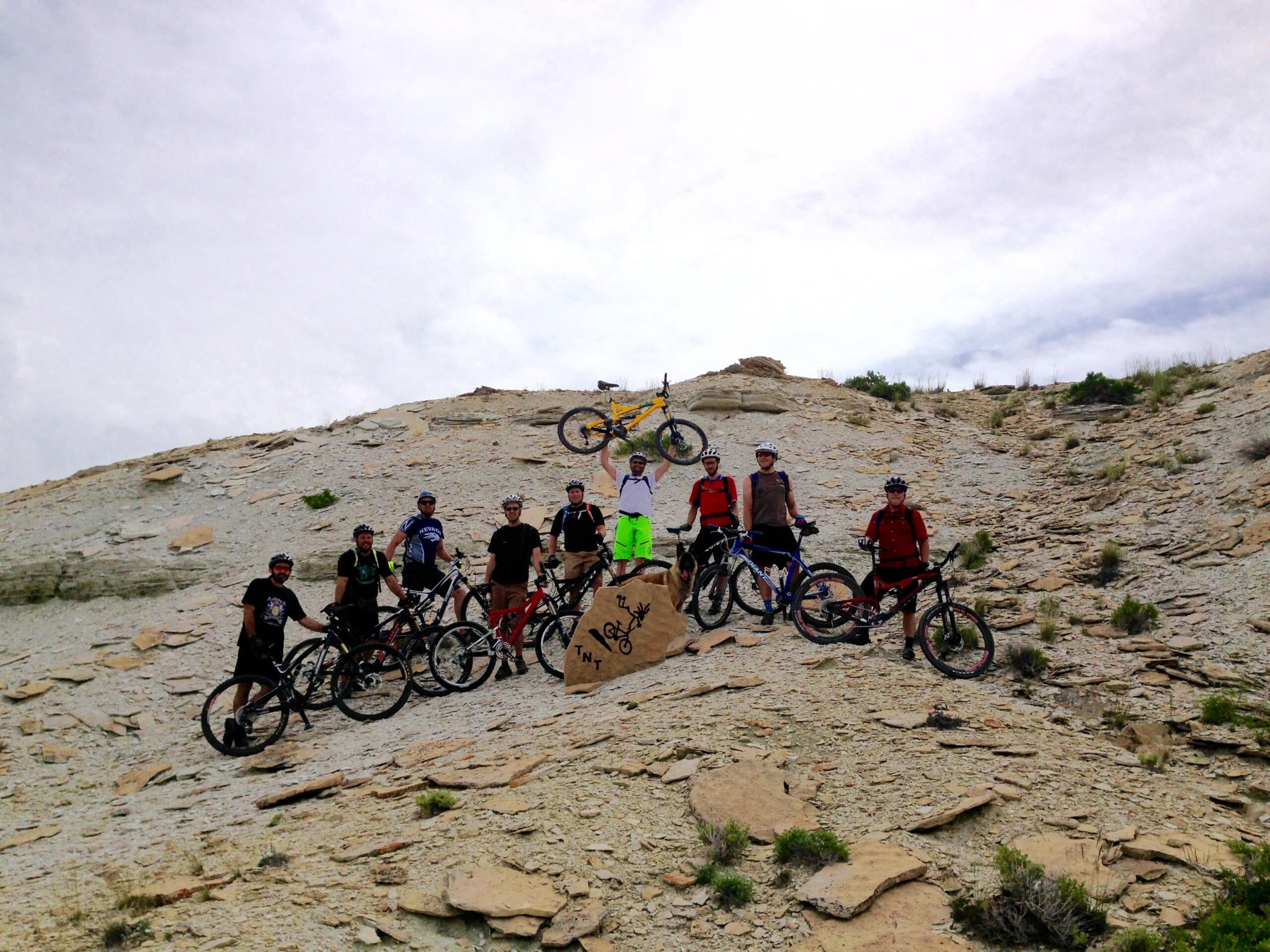 A group of mountain bikers stands on a rocky terrain, posing with their bicycles against a backdrop of a cloudy sky. The team is diverse, wearing helmets and cycling gear, and one individual is holding a bike overhead. A large rock in the foreground is marked with the word "TNT." The scene captures the spirit of outdoor adventure and camaraderie among cyclists. Tnt mountain bike trail.