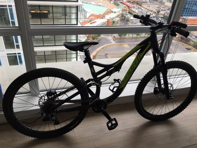 Specialized Stumpjumper FSR 27.5 650B: A black and green mountain bike is leaning against a large window with a view of a parking lot and buildings outside. The bike features thick tires and a sturdy frame, suitable for off-road terrain. The interior space has wooden flooring, and natural light is coming through the window.