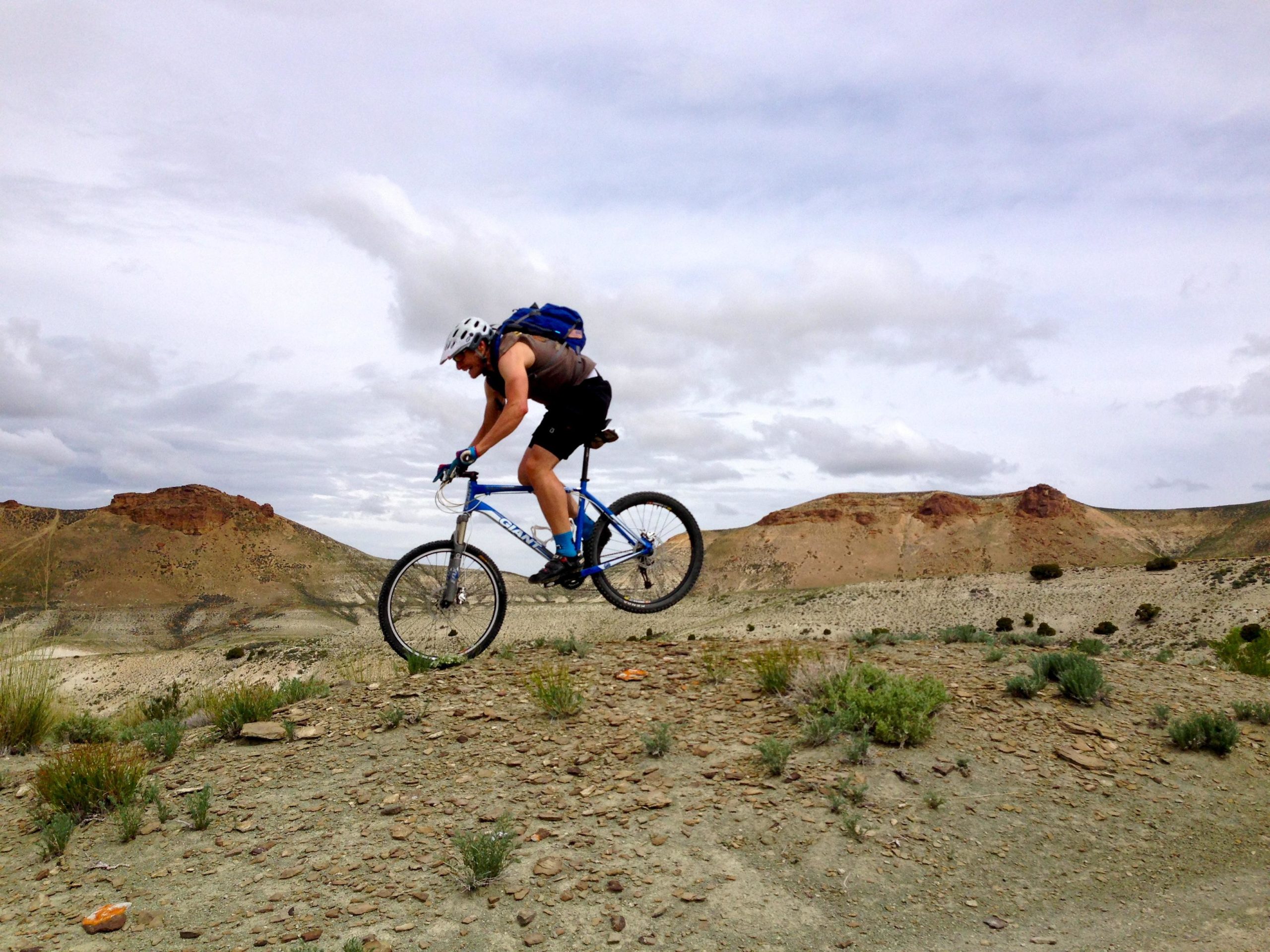 Mountain biker catching air while jumping over a rocky terrain with hills in the background under a cloudy sky. Wilkins Peak Trails mountain bike trail.
