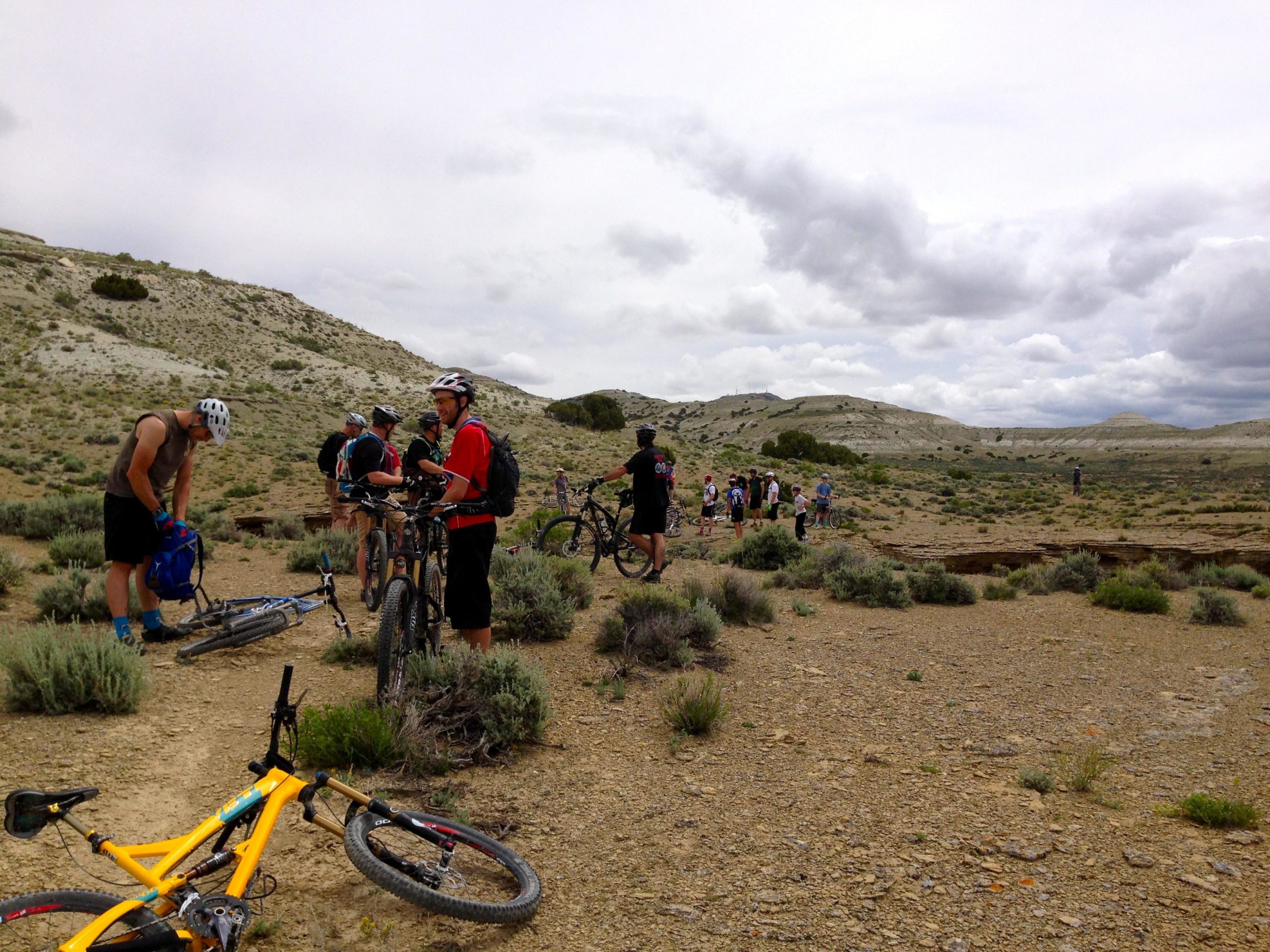 A group of mountain bikers is gathered in a rocky, arid landscape, preparing for a ride. Several bikes are resting on the ground, and riders are wearing helmets and casual sportswear. The background features rolling hills and a cloudy sky, indicating an overcast day. Some riders are examining their gear while others stand nearby, surrounded by shrubs and sparse vegetation. Wilkins Peak Trails mountain bike trail.