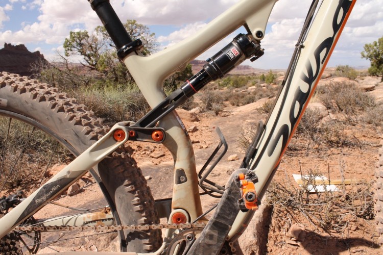 Close-up view of a mountain bike frame featuring suspension components, including shock and linkage, set against a rugged outdoor landscape with rocky terrain and sparse vegetation.