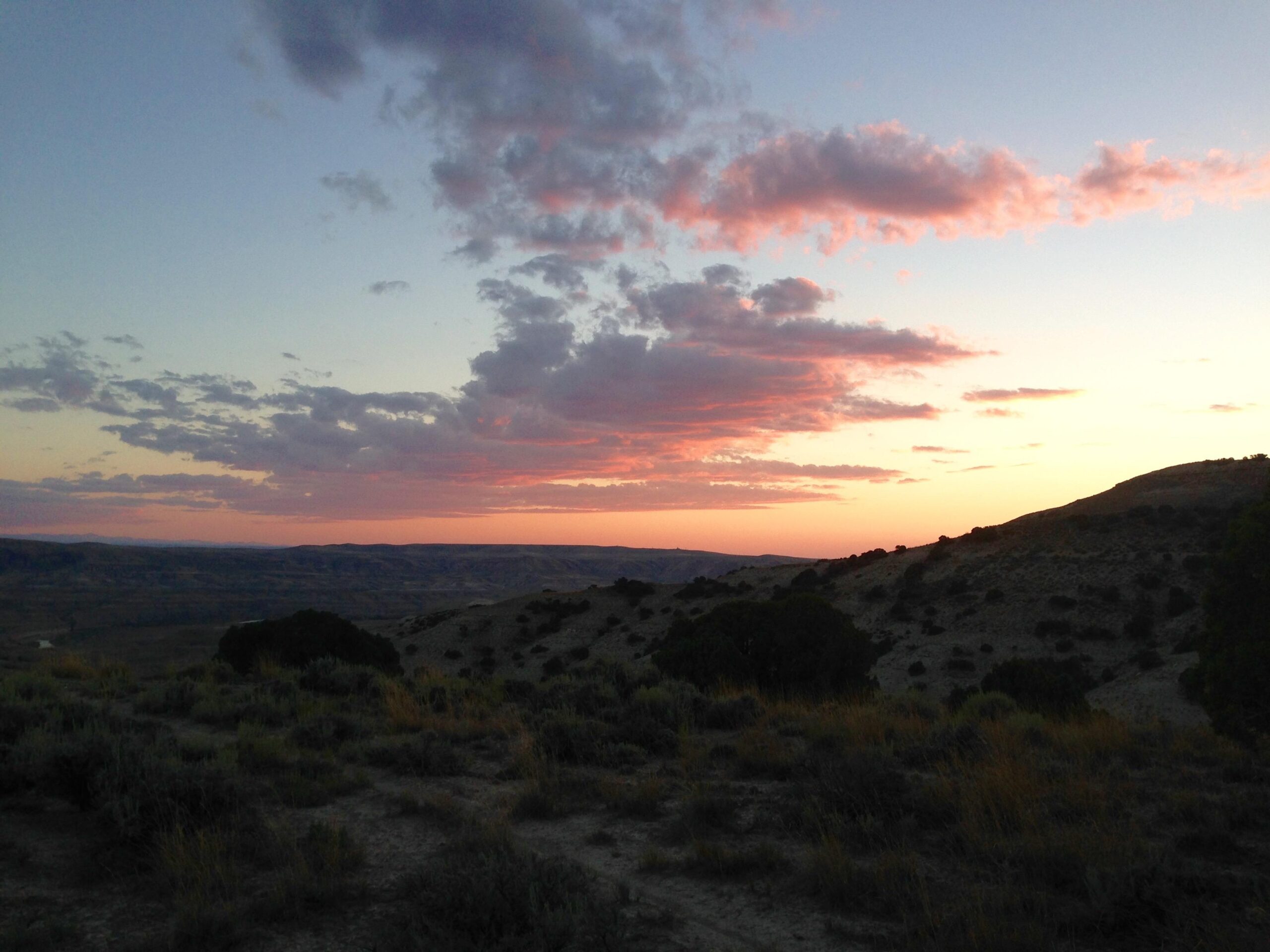 A scenic view of a sunset over rolling hills, featuring a gradient of colors in the sky, with soft pink and purple clouds. The foreground includes sparse vegetation and shrubs, while the horizon displays distant hills and valleys beneath a fading light. Wilkins Peak Trails mountain bike trail.
