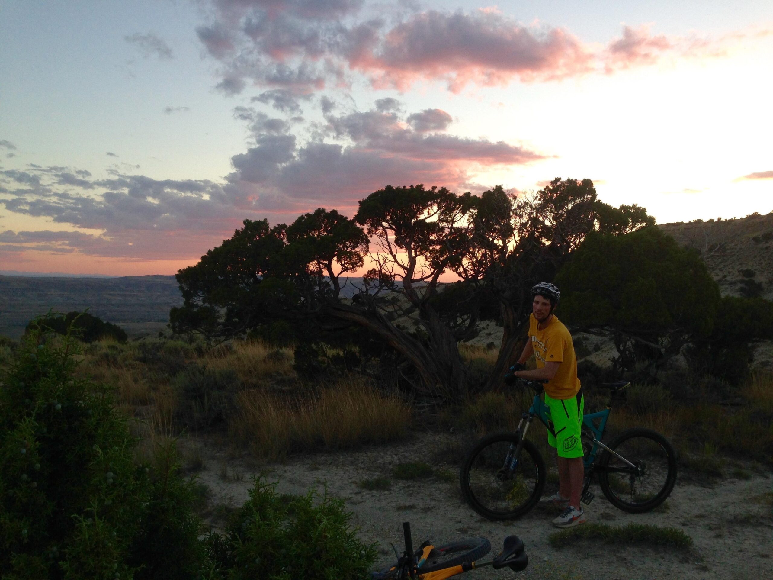 A mountain biker in a bright yellow t-shirt and neon green shorts stands next to his bike on a dirt trail. The scene features a beautiful sunset with pink and purple hues in the sky, surrounded by lightly wooded terrain and distant hills. Wilkins Peak Trails mountain bike trail.