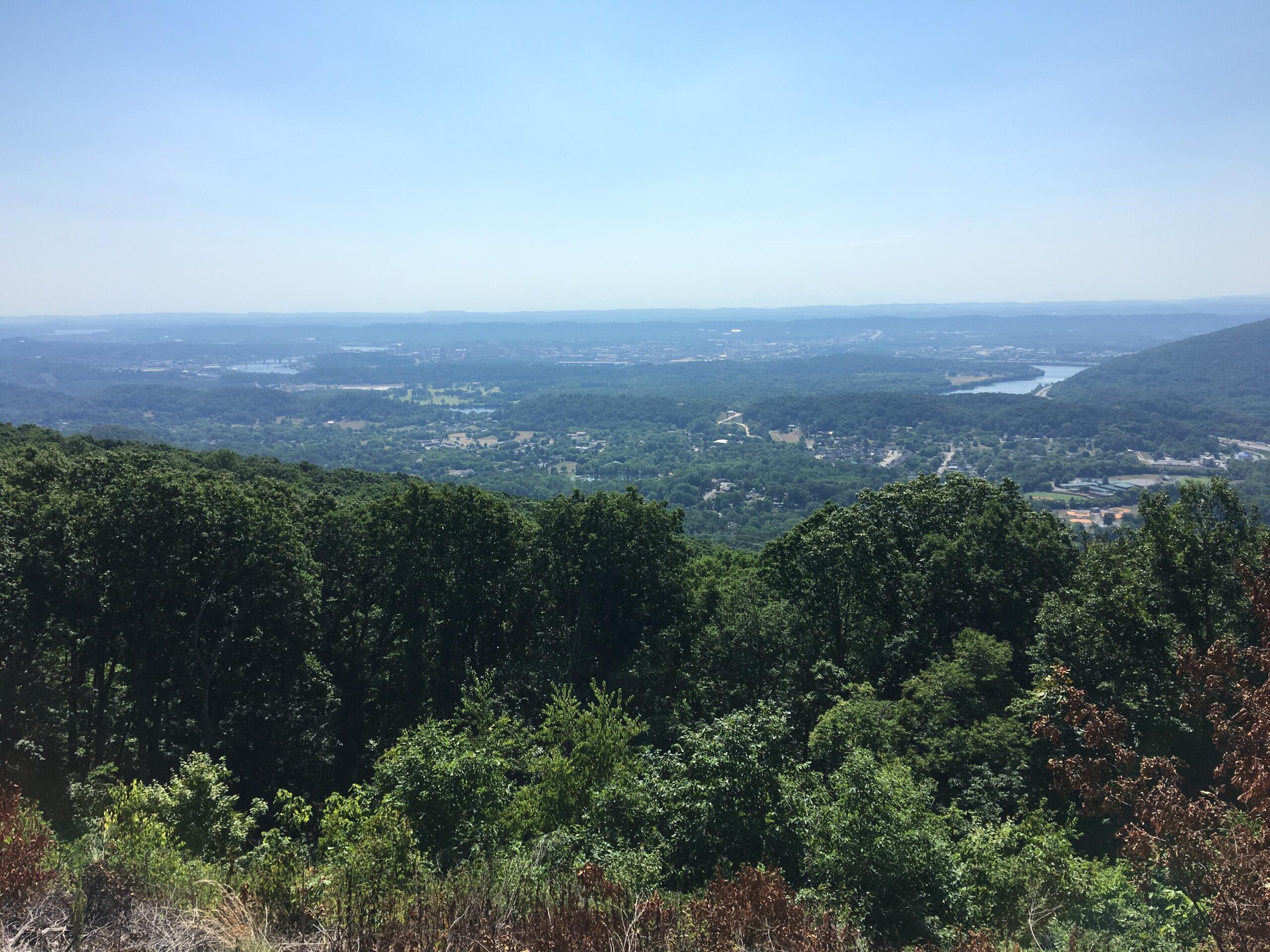 A panoramic view of a lush, green valley from a mountain overlook, featuring dense trees in the foreground and rolling hills in the background under a clear blue sky. A river meanders through the valley, with small towns and fields visible in the distance. Raccoon Mountain Trail Network mountain bike trail.
