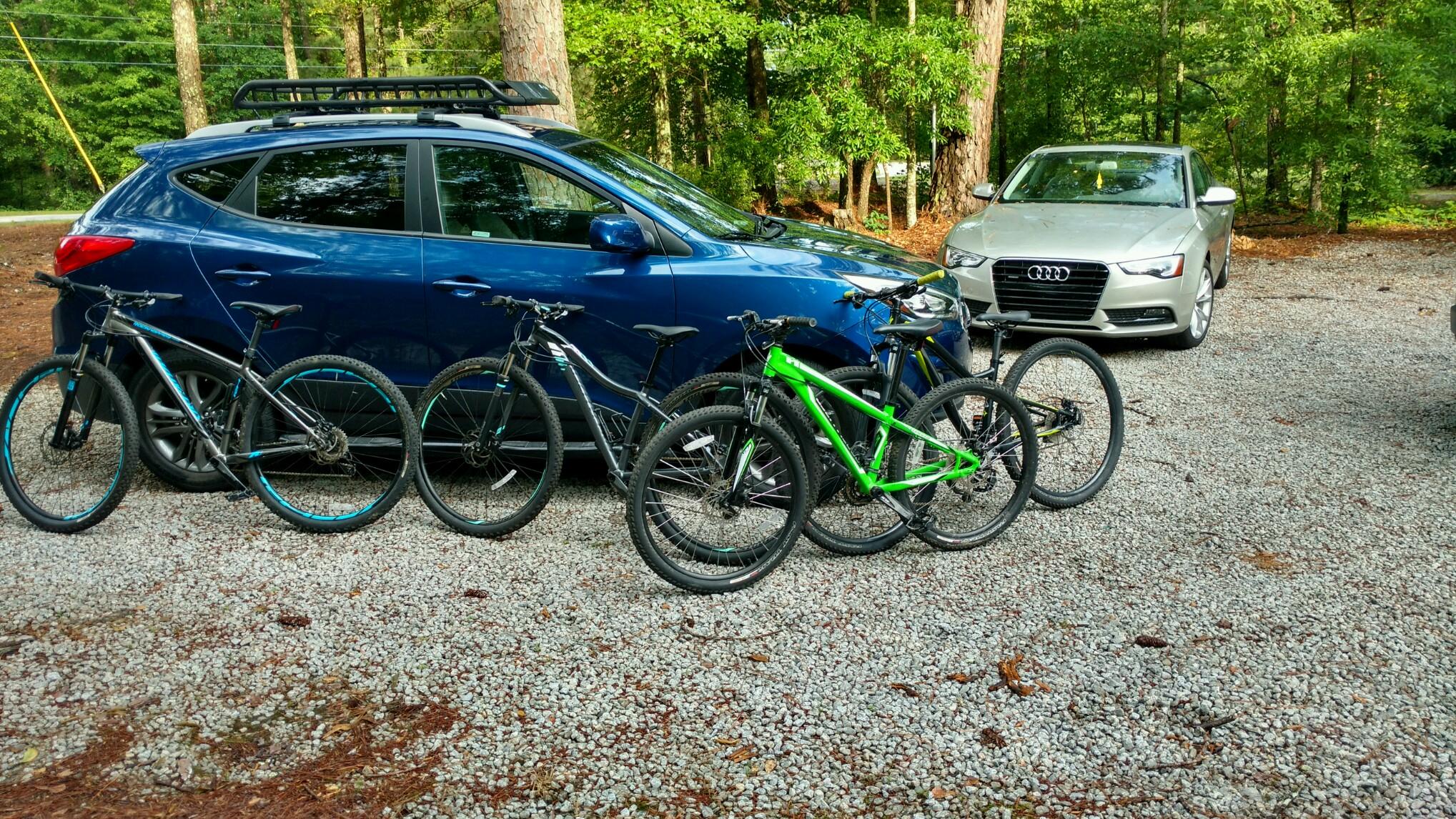 Specialized hardrock: A blue car parked on a gravel surface with several mountain bikes arranged in front of it, including a bright green bike. A silver car is visible in the background, surrounded by trees.