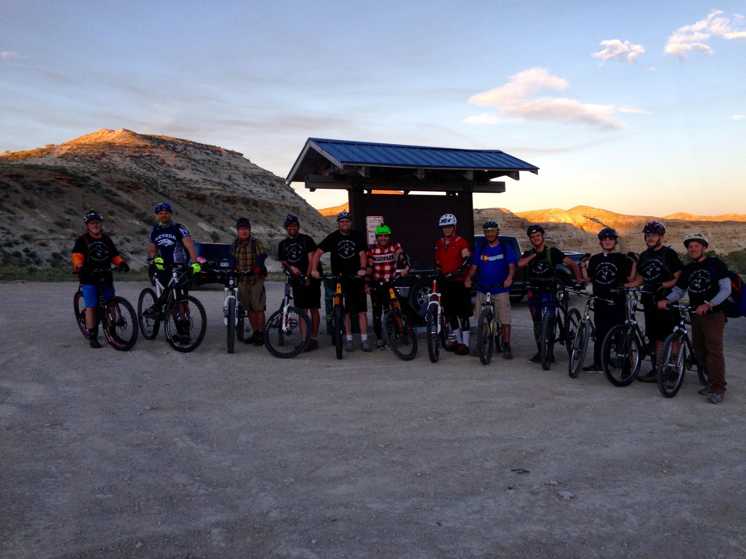 A group of ten people, dressed in casual cycling attire and wearing helmets, are gathered near a small pavilion in a desert landscape. They are all positioned beside their mountain bikes, with sunlit hills in the background and a clear sky above, indicating a late afternoon setting. The group displays a mix of colors and styles in their clothing, suggesting a camaraderie among fellow cyclists. Wilkins Peak Trails mountain bike trail.