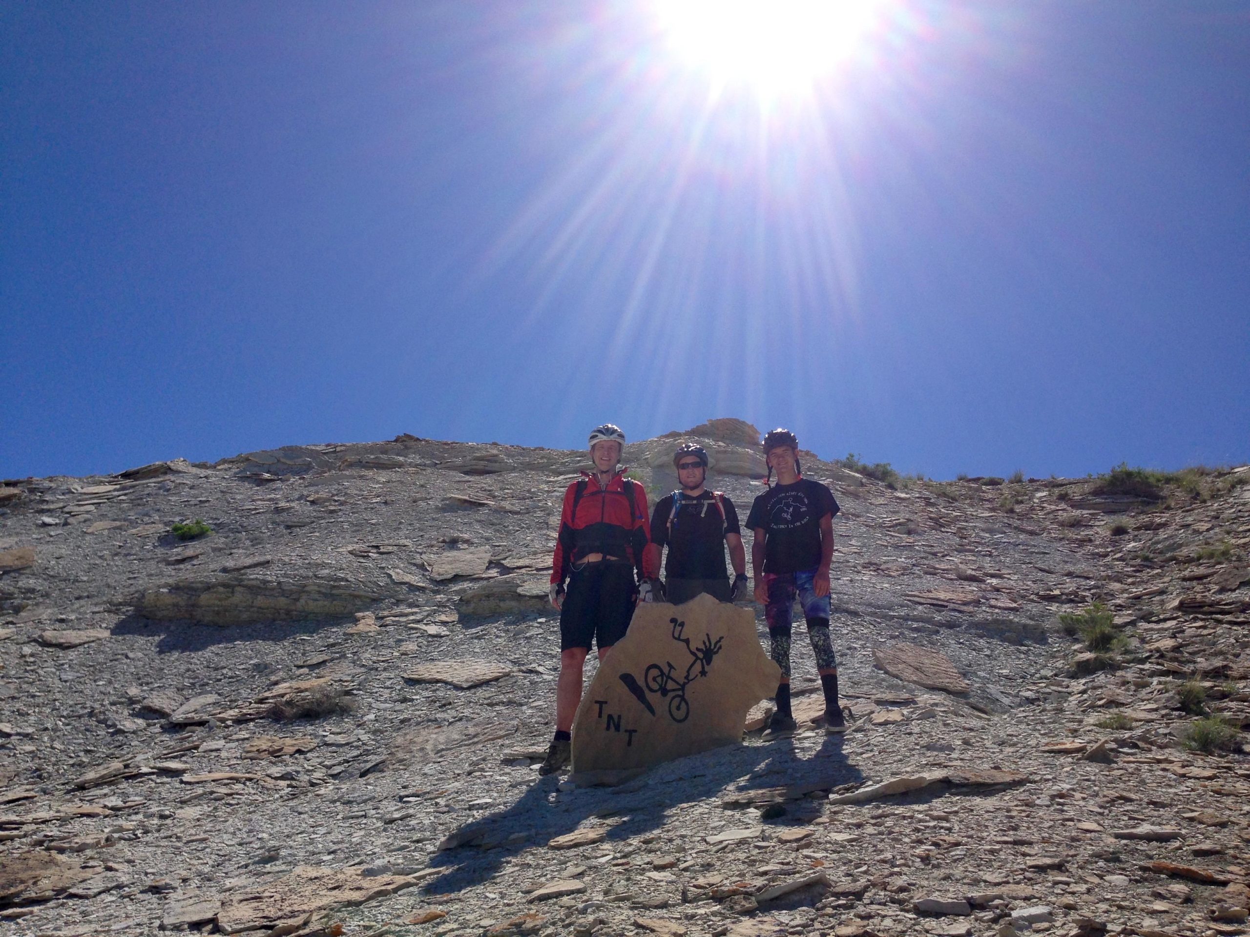 Three cyclists stand on a rocky slope under a clear blue sky, with the sun shining brightly above them. They are wearing helmets and riding gear, with a prominent rock featuring a graffiti-style design labeled "TNT" nearby. Wilkins Peak Trails mountain bike trail.