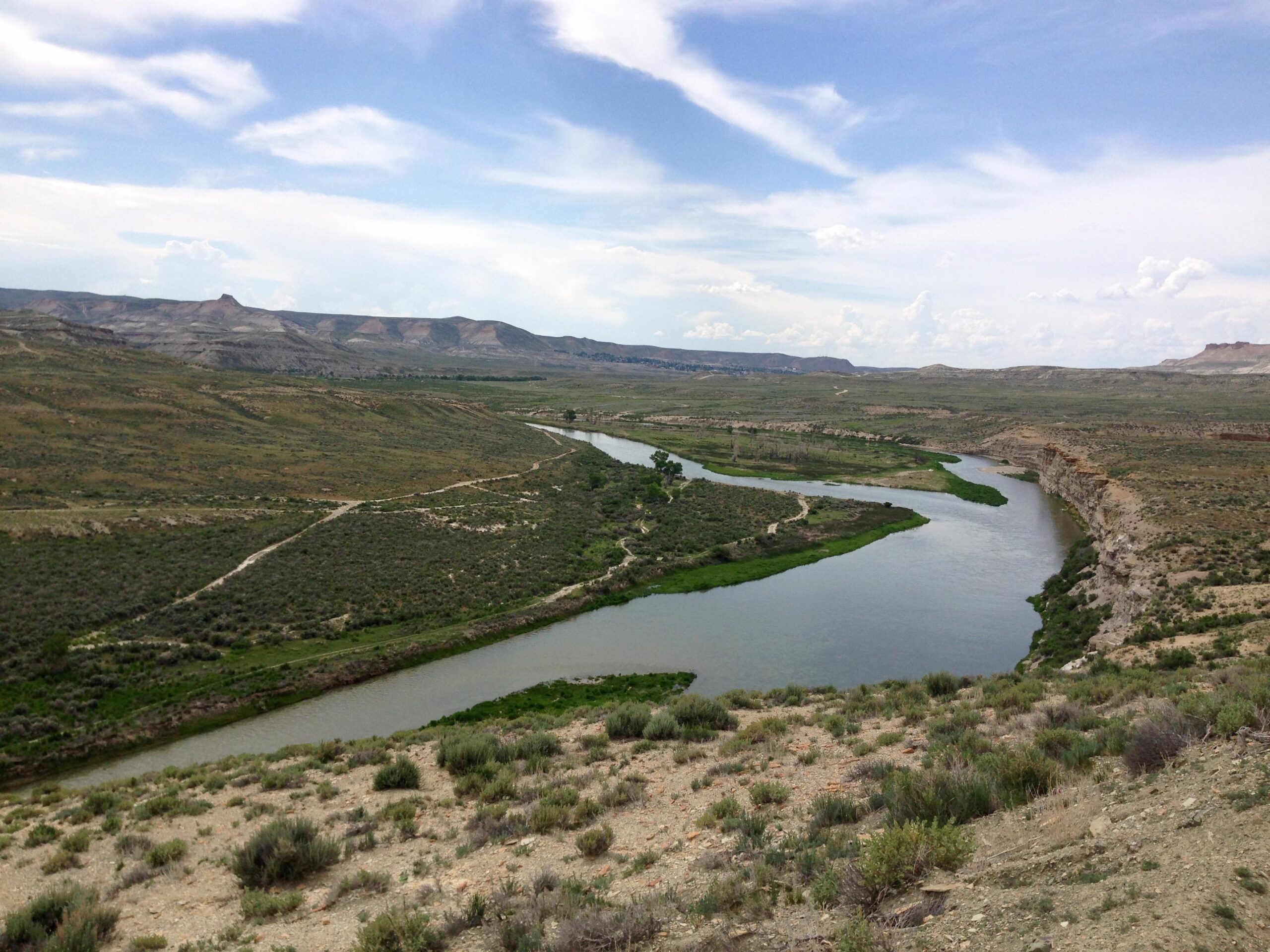 A scenic landscape view featuring a winding river surrounded by lush green vegetation, with rolling hills and rocky cliffs in the background under a partly cloudy sky. The scene captures the natural beauty of a remote, open area, with hills rising in the distance. Wilkins Peak Trails mountain bike trail.
