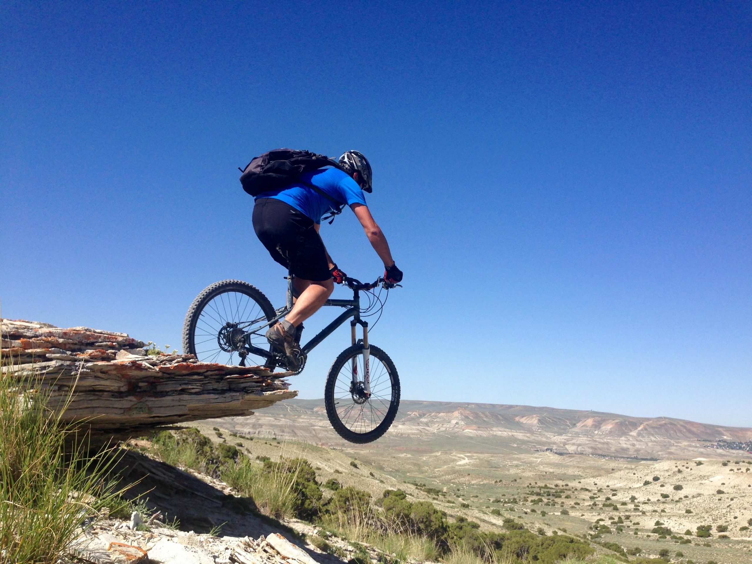 A mountain biker in a blue shirt and black shorts is balancing on a ledge with a steep drop-off, overlooking a vast landscape with rolling hills and a clear blue sky. The biker is wearing a helmet and a backpack, focused on the terrain ahead. Brent & Mikes' Trail mountain bike trail.